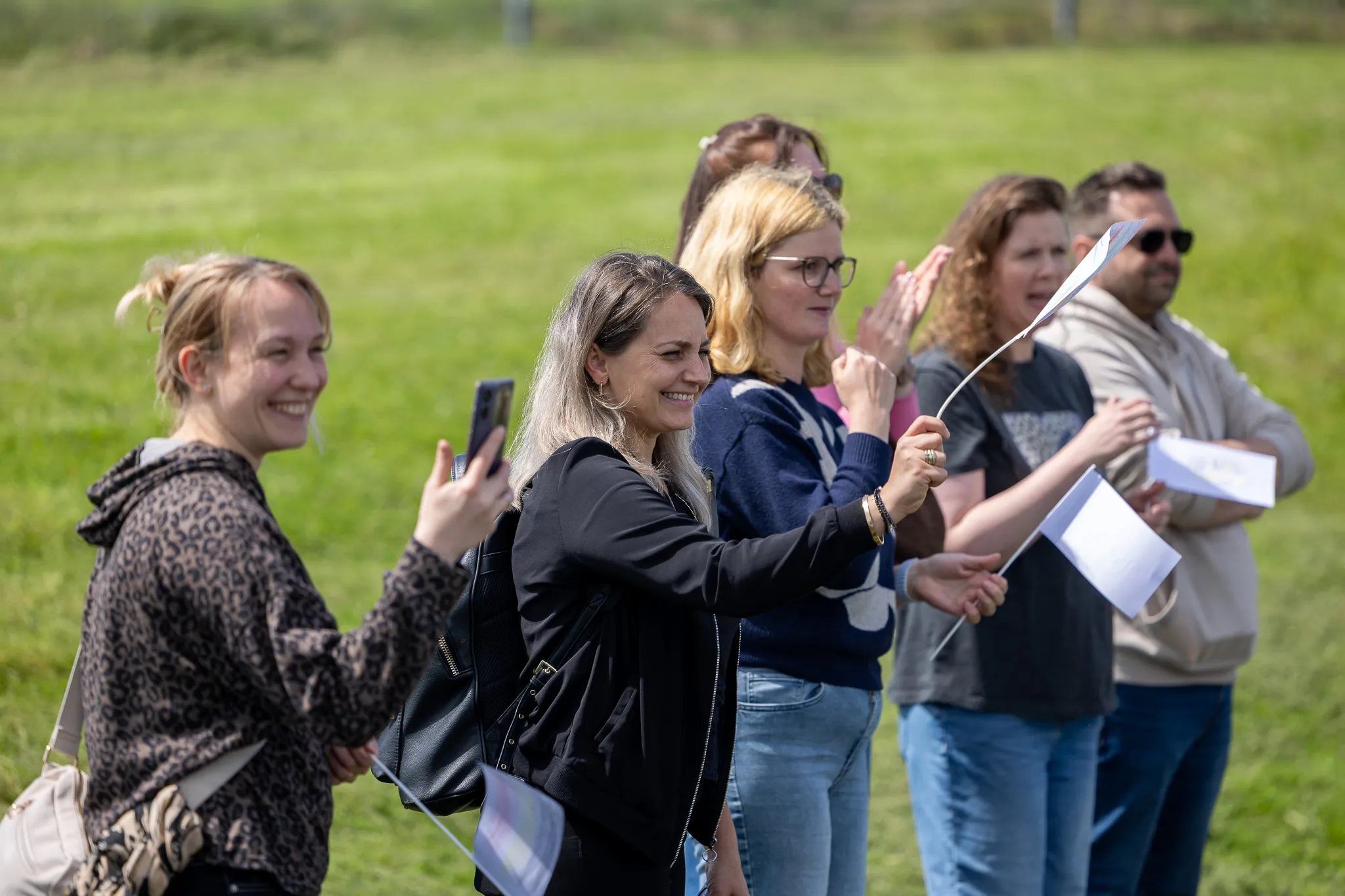 Eine Gruppe von Menschen steht auf einer grünen Wiese und verfolgt ein Fußballturnier. Einige halten Papiere oder Smartphones in der Hand und lächeln.