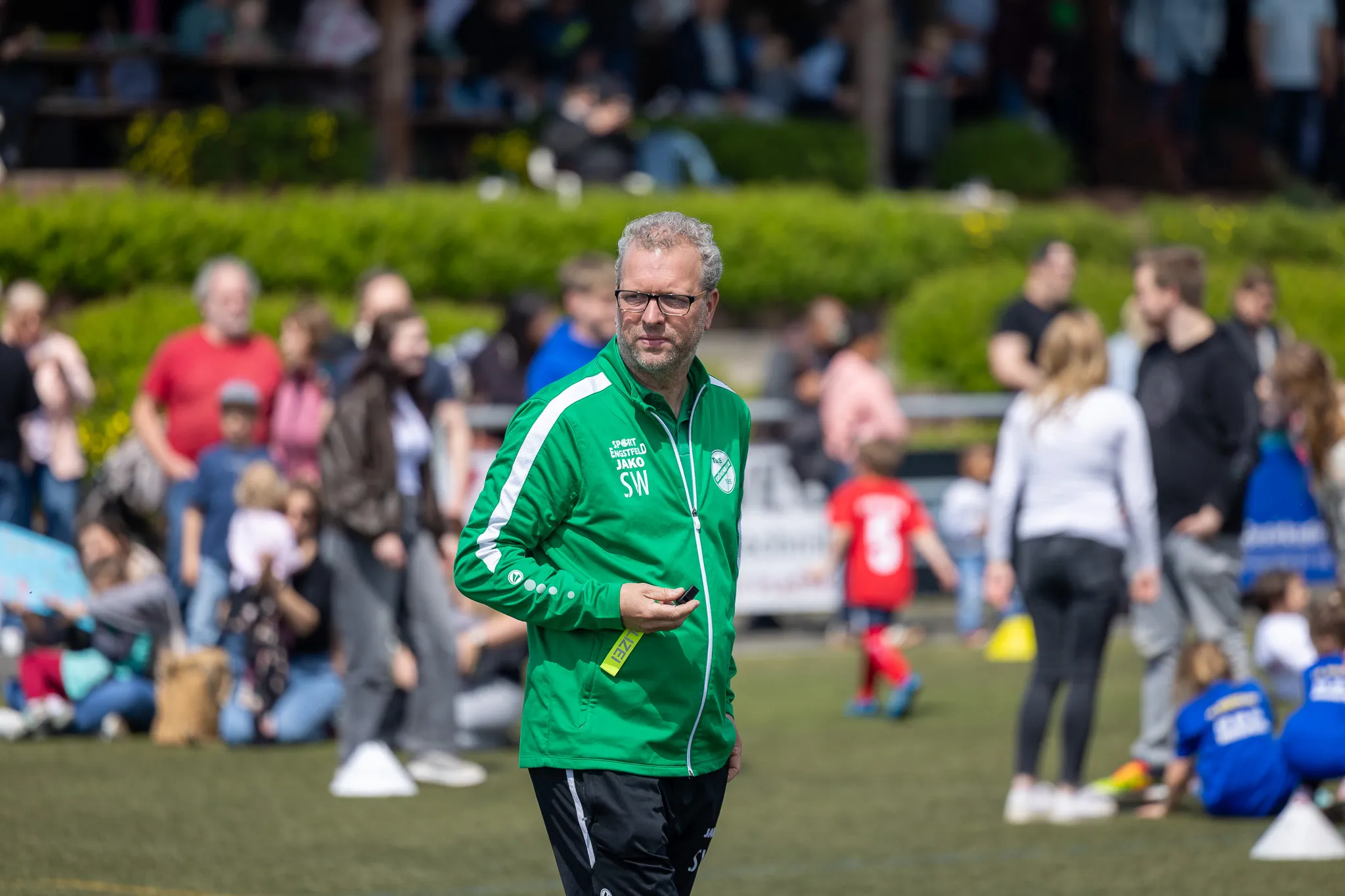 Ein Mann mit Brille in grüner Trainingsjacke steht am Spielfeldrand und hält ein Smartphone in der Hand, während im Hintergrund Kinder Fußball spielen und Zuschauer zusehen.