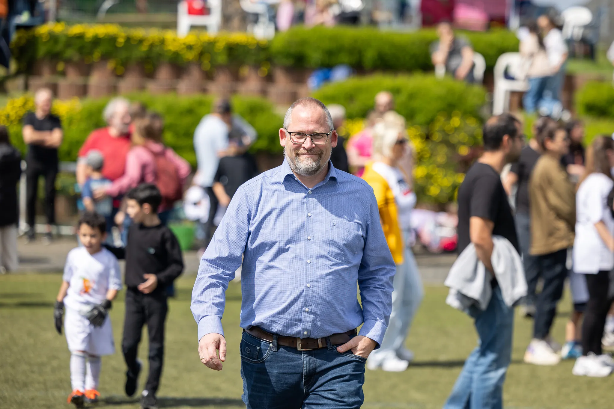 Ein Mann mit Bart und Brille geht lächelnd durch eine Menschenmenge auf einer Grünfläche. Im Hintergrund sind weitere Personen und gelbe Blumen zu sehen.