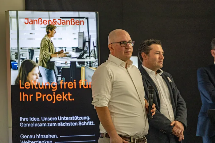Ein Mann mit Brille und weißem Hemd hält ein Mikrofon vor einem Plakat der Firma Janßen&Janßen.
