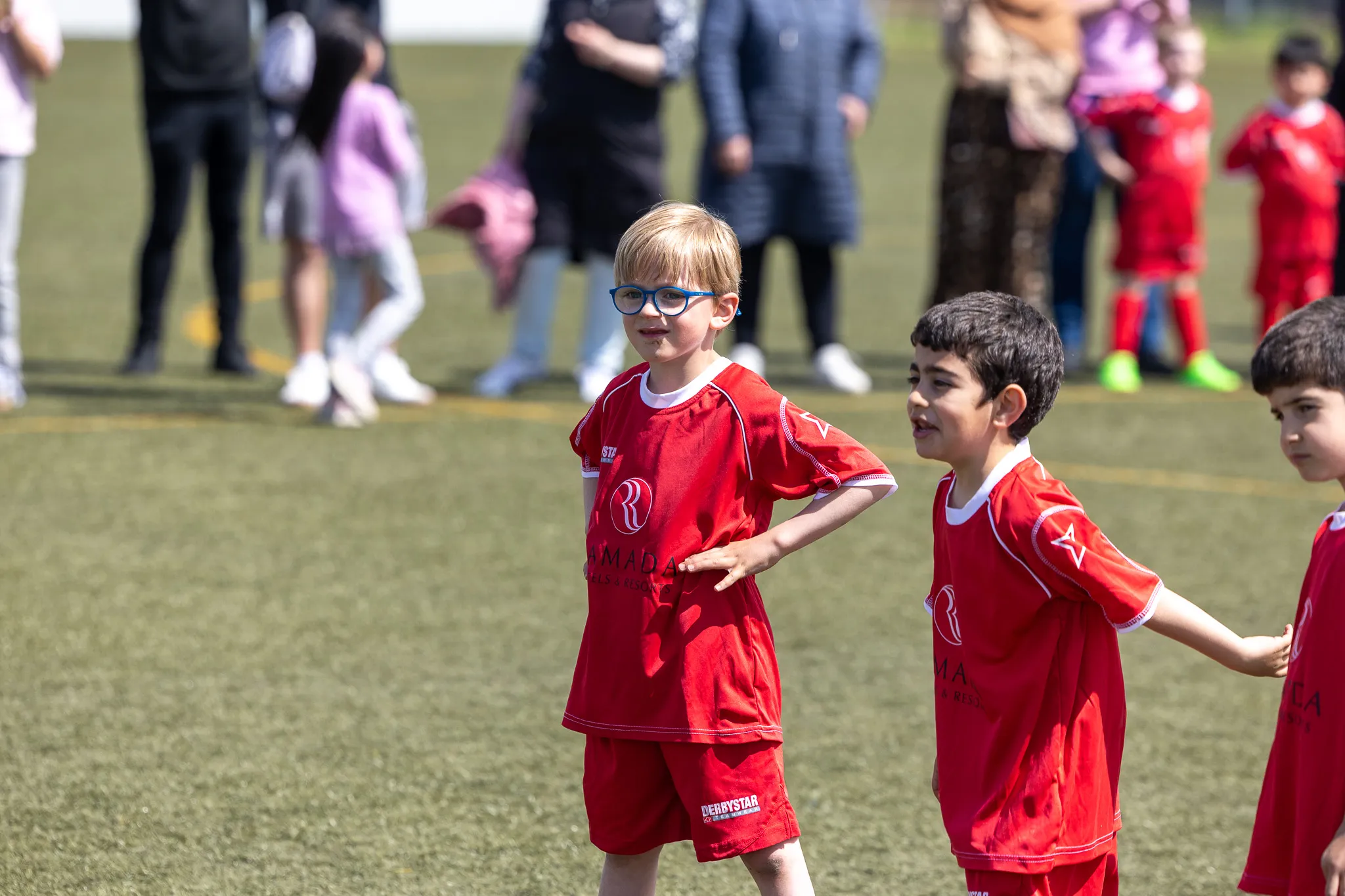 Junge Fußballspieler in roten Trikots stehen auf einem grünen Kunstrasenplatz, während im Hintergrund Zuschauer stehen.