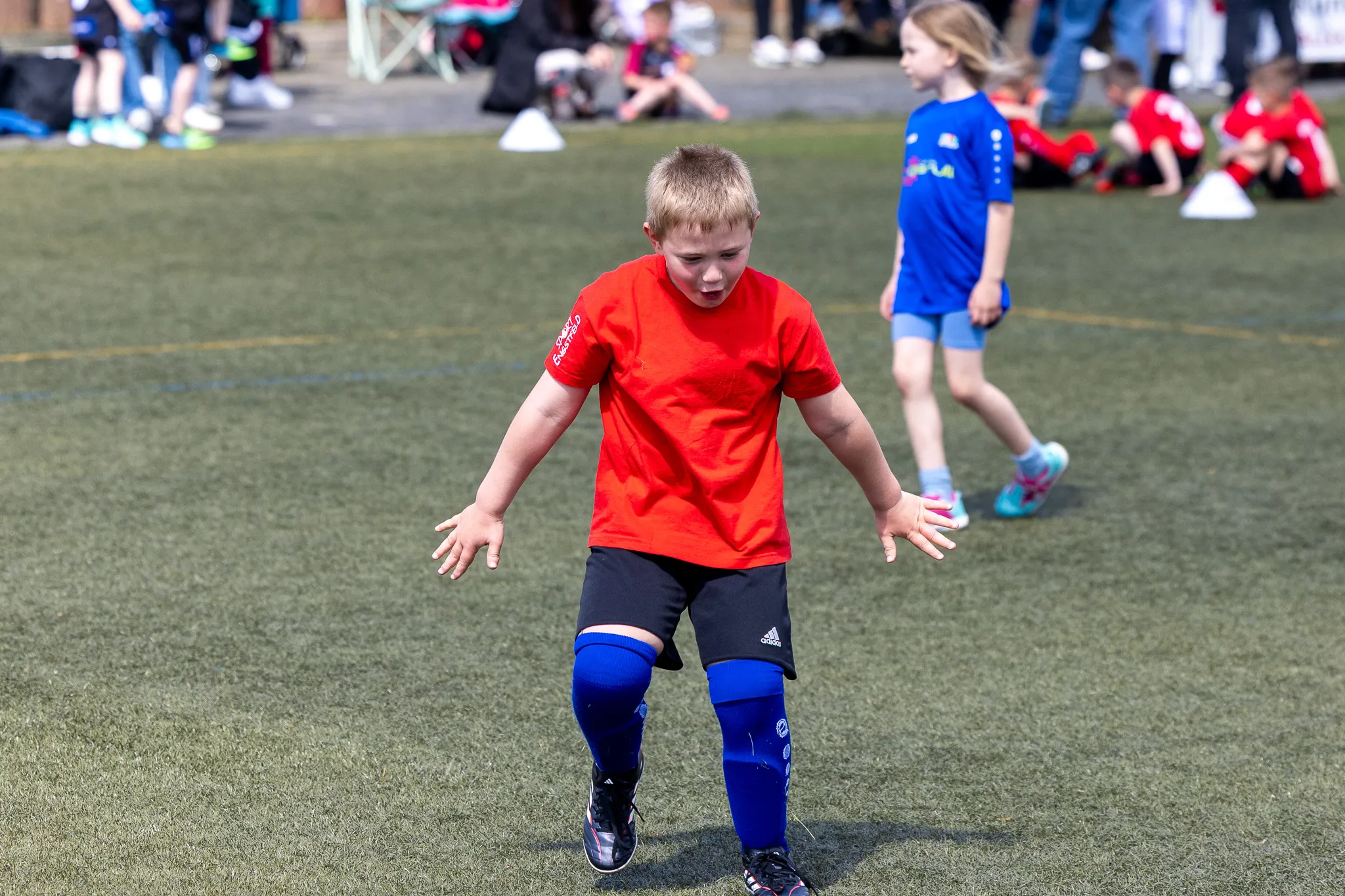 Ein Junge mit Down-Syndrom in rotem T-Shirt und blauen Sportsocken rennt auf einem grünen Kunstrasenplatz, während im Hintergrund andere Kinder spielen und Zuschauer zusehen.