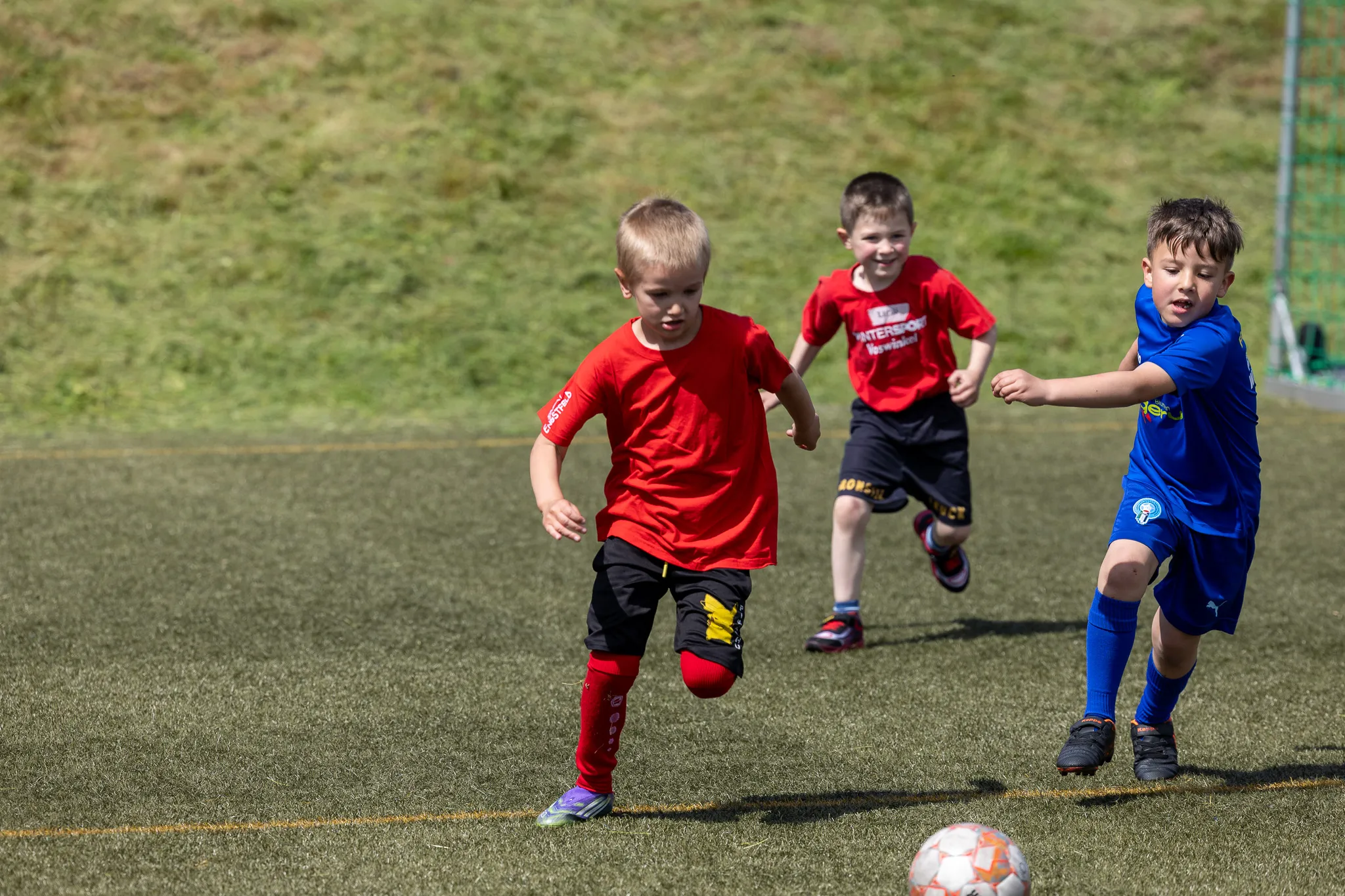 Drei Kinder spielen Fußball auf einer grünen Wiese. Ein Kind in Rot dribbelt den Ball, während zwei weitere Kinder versuchen, ihm den Ball abzunehmen.