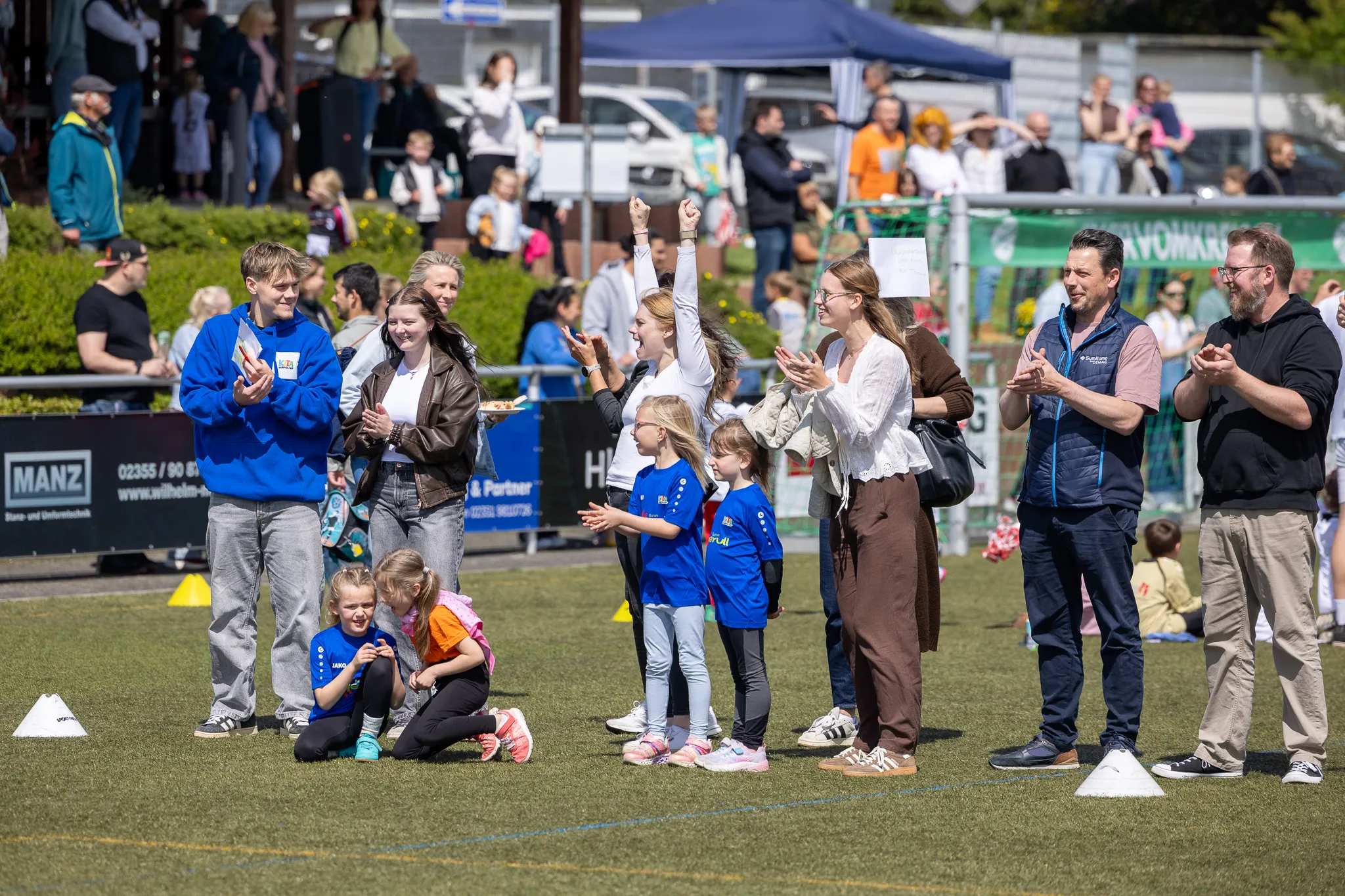 Zuschauer klatschen und jubeln bei einem Fußballturnier für Kinder am Kreisch. Im Vordergrund stehen Eltern und Betreuer, während Kinder im Hintergrund spielen.