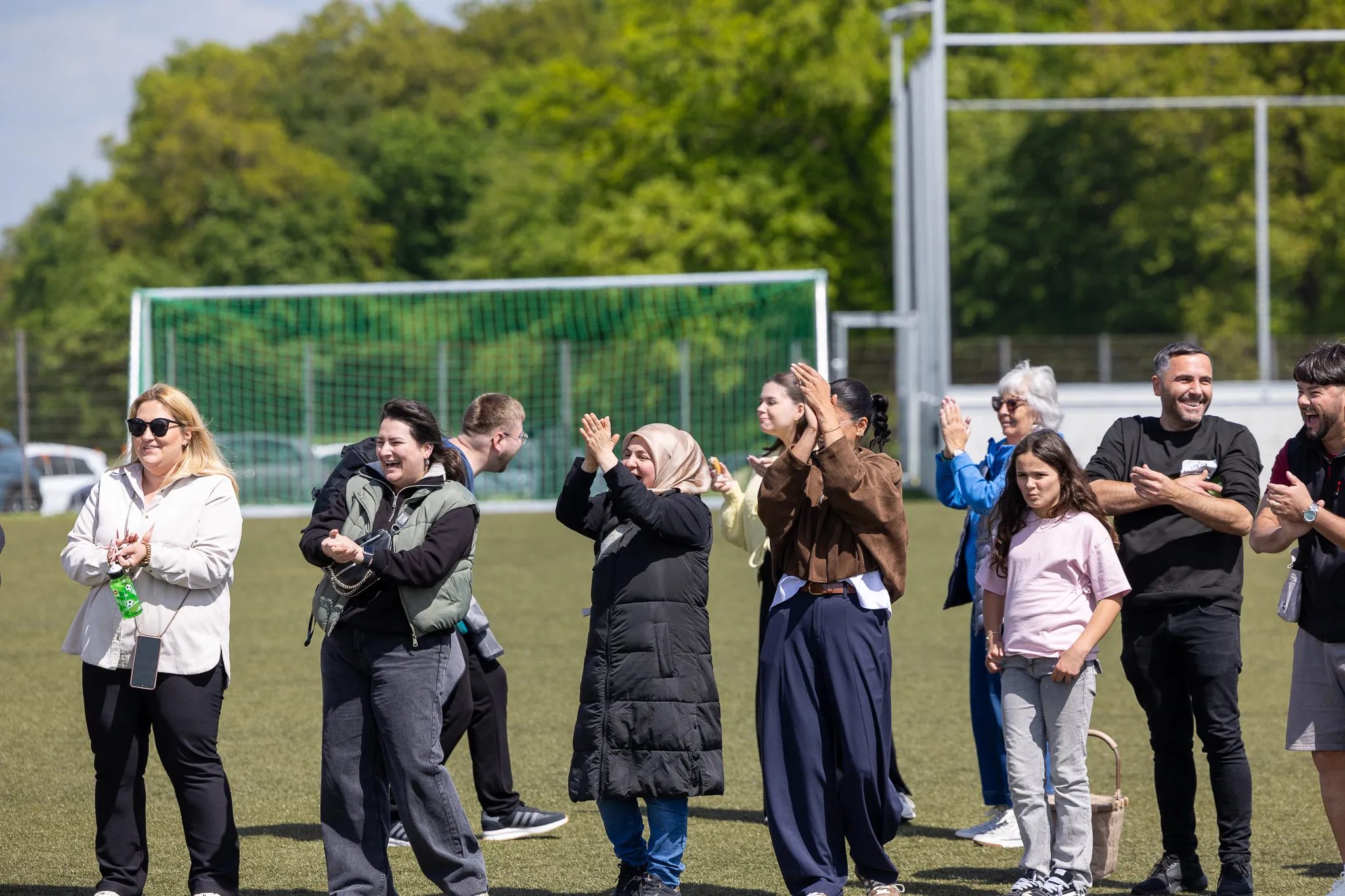 Zuschauer klatschen bei einem Fußballturnier für Kinder auf einem Kunstrasenplatz. Im Hintergrund ein Fußballtor und Bäume.