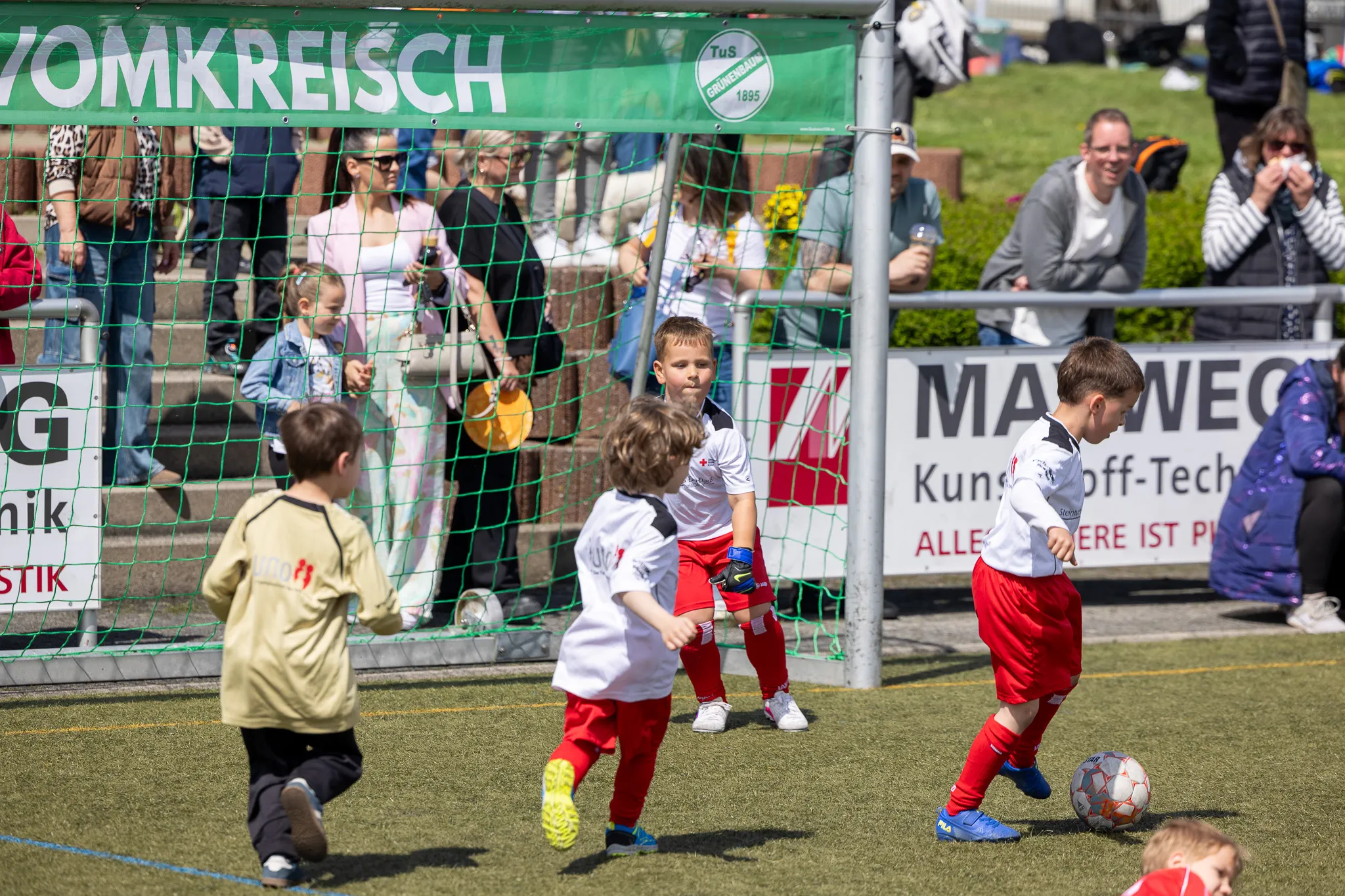 Junge Fußballspieler in roten und weißen Trikots kämpfen um den Ball vor einem Tor, während Zuschauer im Hintergrund zusehen.