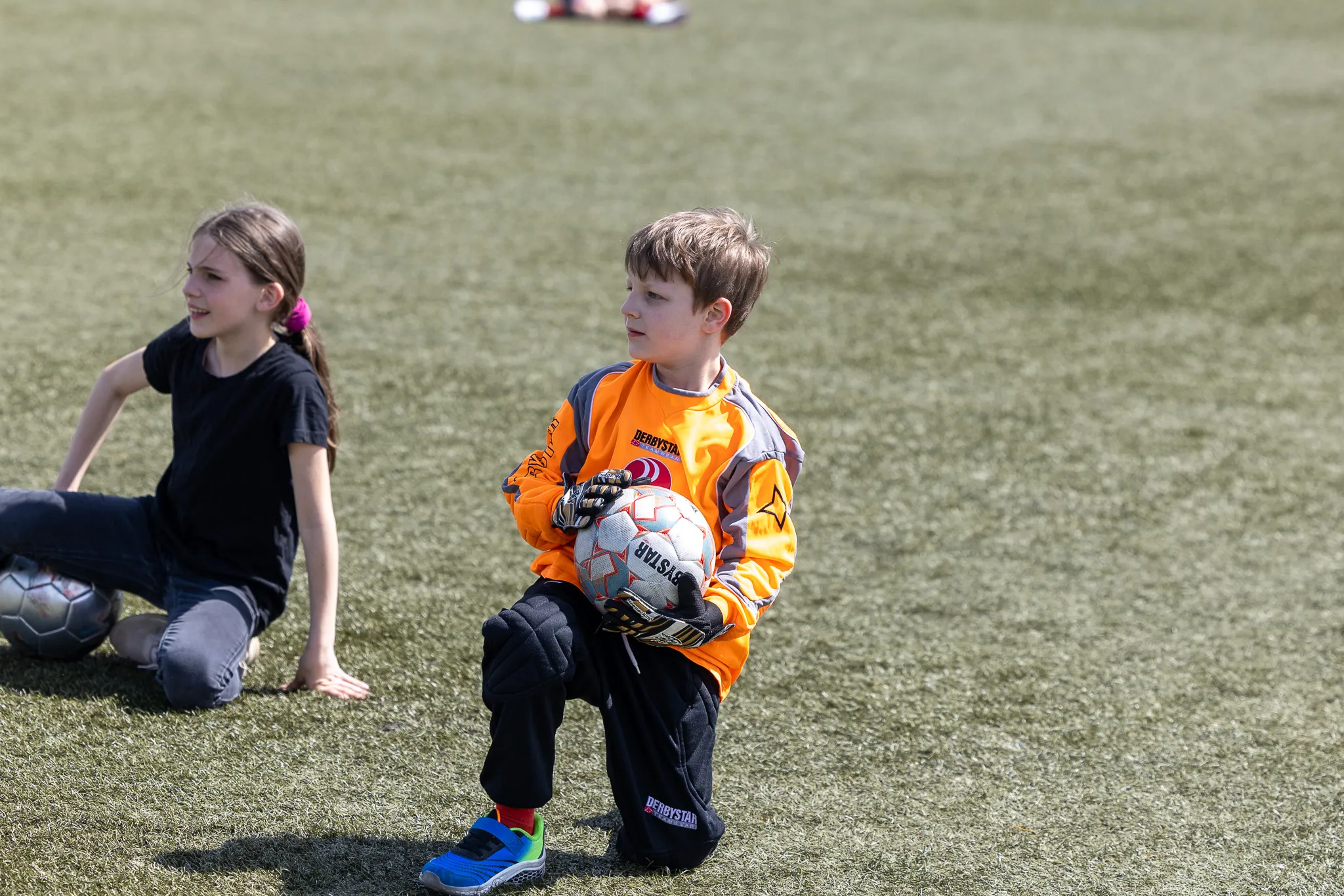 Junge in Torwartmontur hält Fußball, während ein Mädchen im Hintergrund sitzt. Im Hintergrund ist ein Fußballfeld zu sehen.