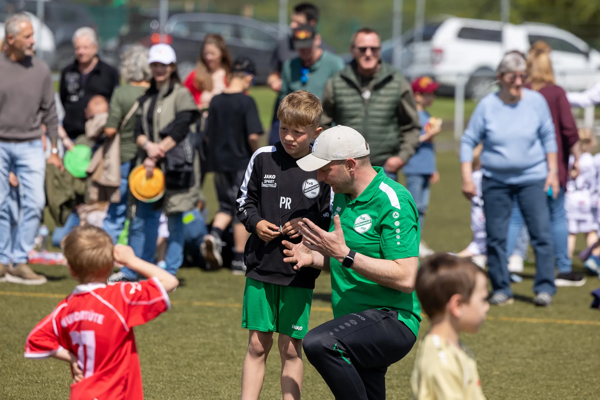 Ein Fußballtrainer kniet vor einem jungen Spieler, um ihn zu motivieren, während im Hintergrund Zuschauer am Spielfeldrand stehen.