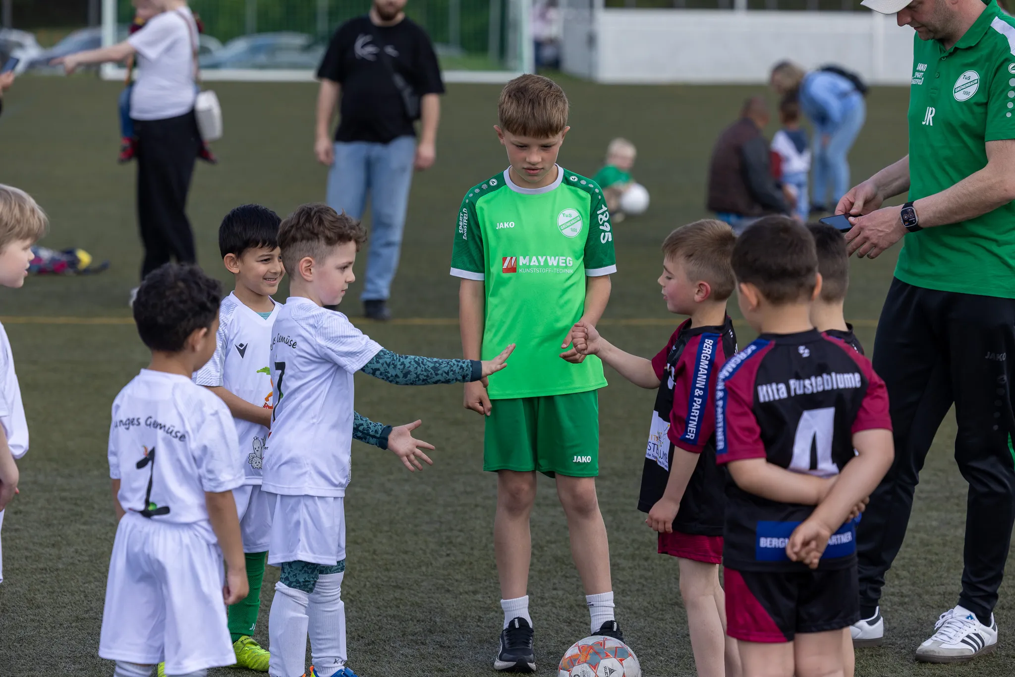 Zwei Fußballmannschaften mit Kindern im Alter von etwa 8 bis 10 Jahren geben sich vor dem Spiel die Hände. Im Hintergrund stehen weitere Kinder und Betreuer am Spielfeldrand.