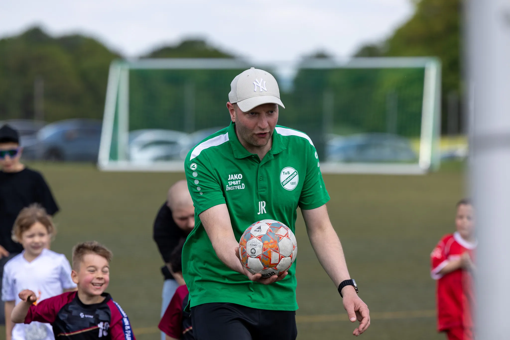 Ein Mann in grünem Trainingsanzug hält einen Fußball und blickt auf Kinder, die ihn umringen. Im Hintergrund ein Fußballtor und weitere Personen auf einem Rasenplatz.