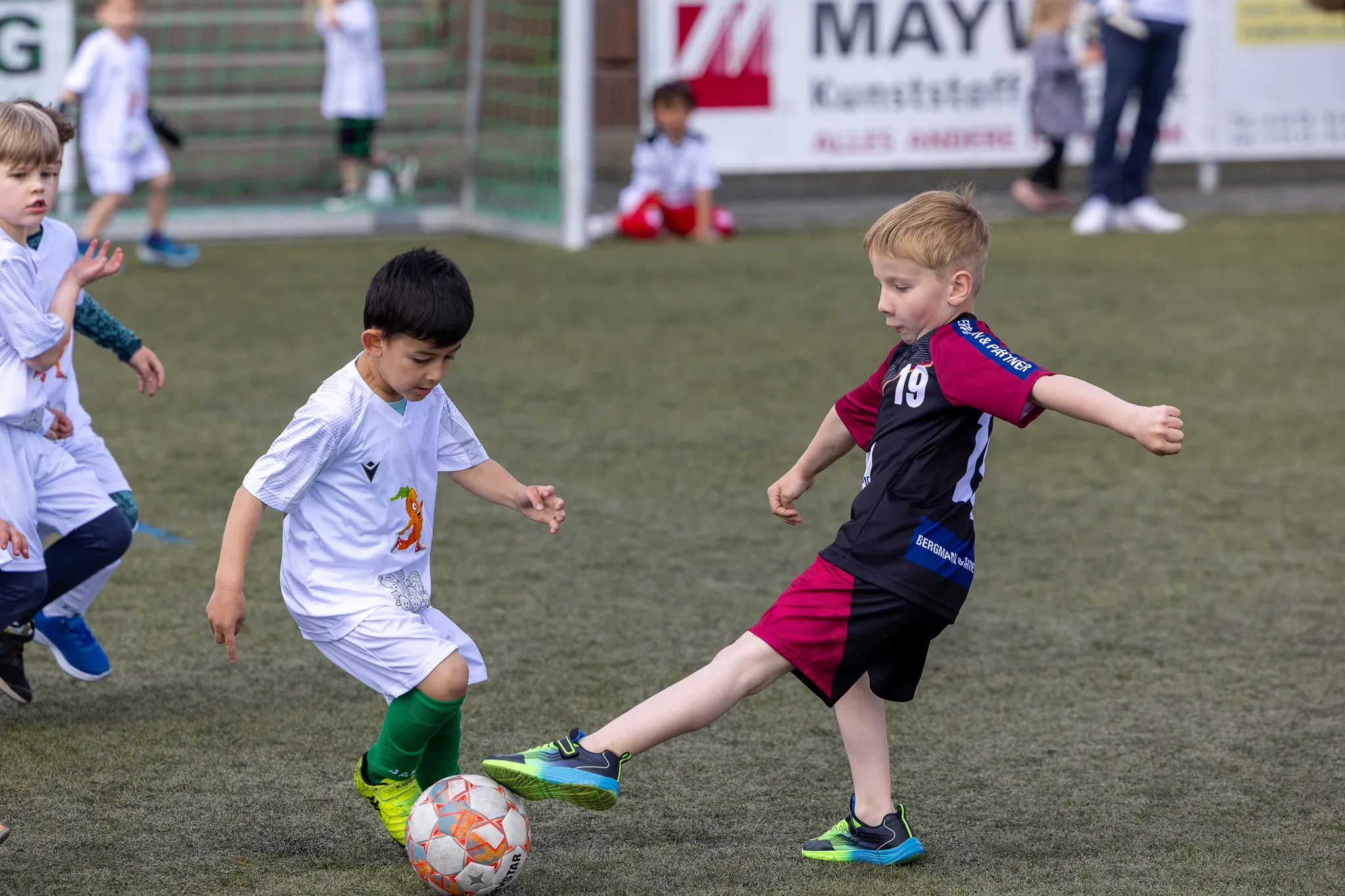 Zwei junge Fußballspieler treten auf einem Kunstrasenfeld an, während andere Kinder im Hintergrund zusehen. Der Spieler links trägt ein grünes Trikot, der andere ein dunkelrotes mit schwarzen Akzenten.