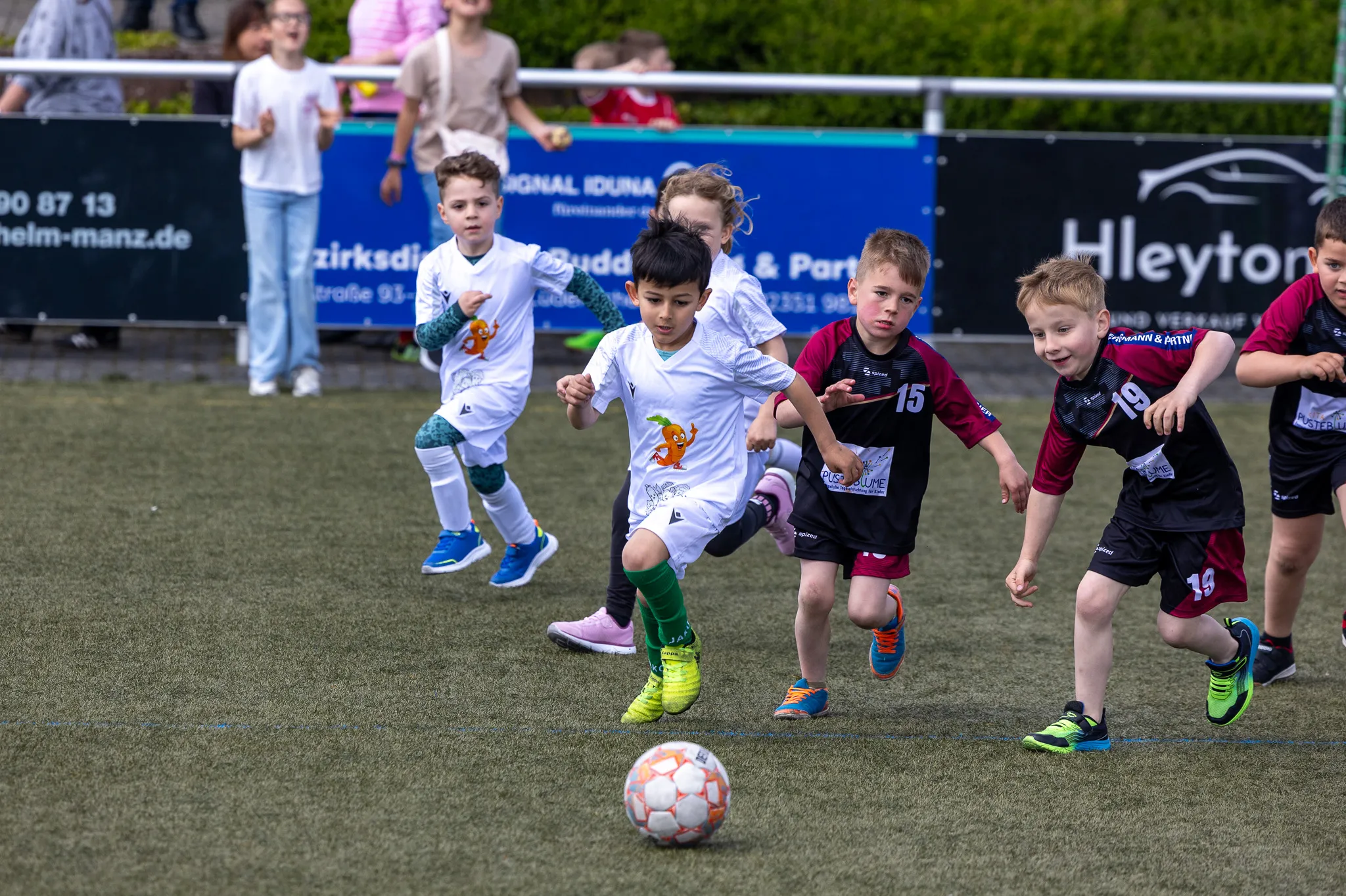 Kinder spielen Fußball auf einem Kunstrasenplatz, einige rennen dem Ball hinterher, andere versuchen ihn zu erobern. Im Hintergrund sind Zuschauer und Werbebanner zu sehen.