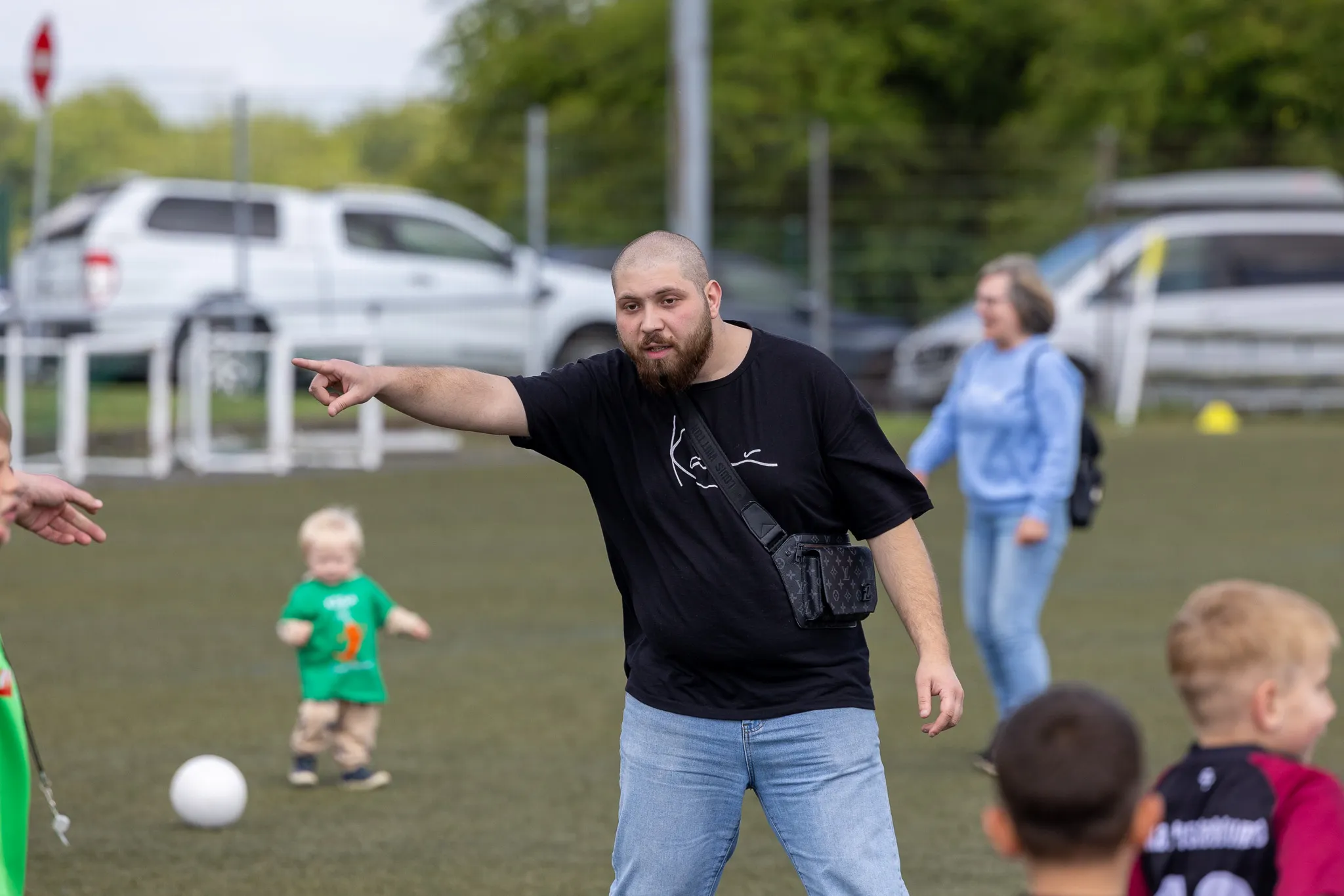Ein Mann mit Bart zeigt mit ausgestrecktem Arm auf ein Kind, das mit einem Fußball spielt. Im Hintergrund sind weitere Kinder und Erwachsene sowie geparkte Autos zu sehen.