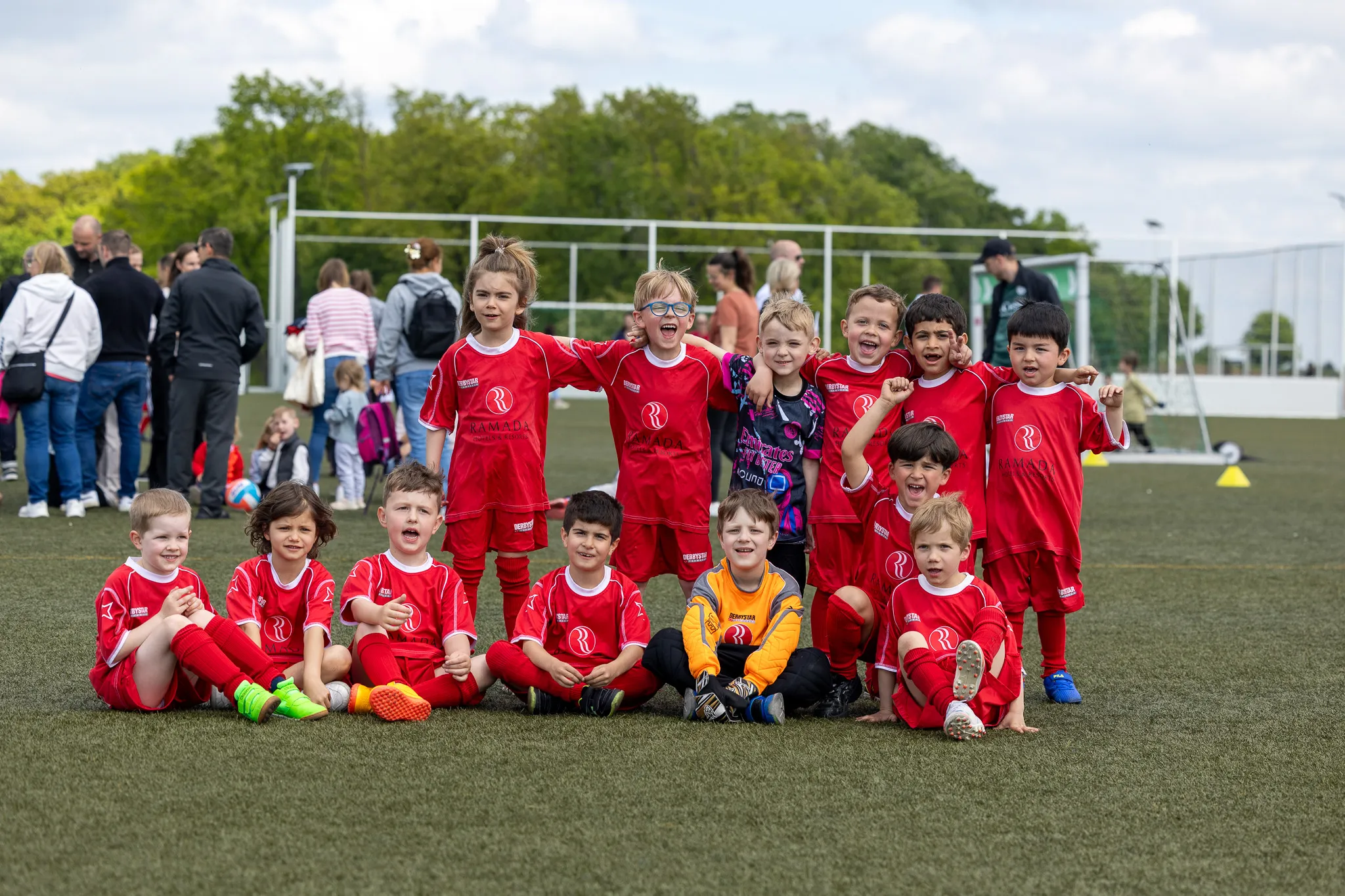 Eine Gruppe von Kindern im Fußballtrikot sitzt und steht auf einem grünen Rasenfeld, einige lächeln und jubeln in die Kamera. Im Hintergrund stehen Zuschauer am Spielfeldrand.