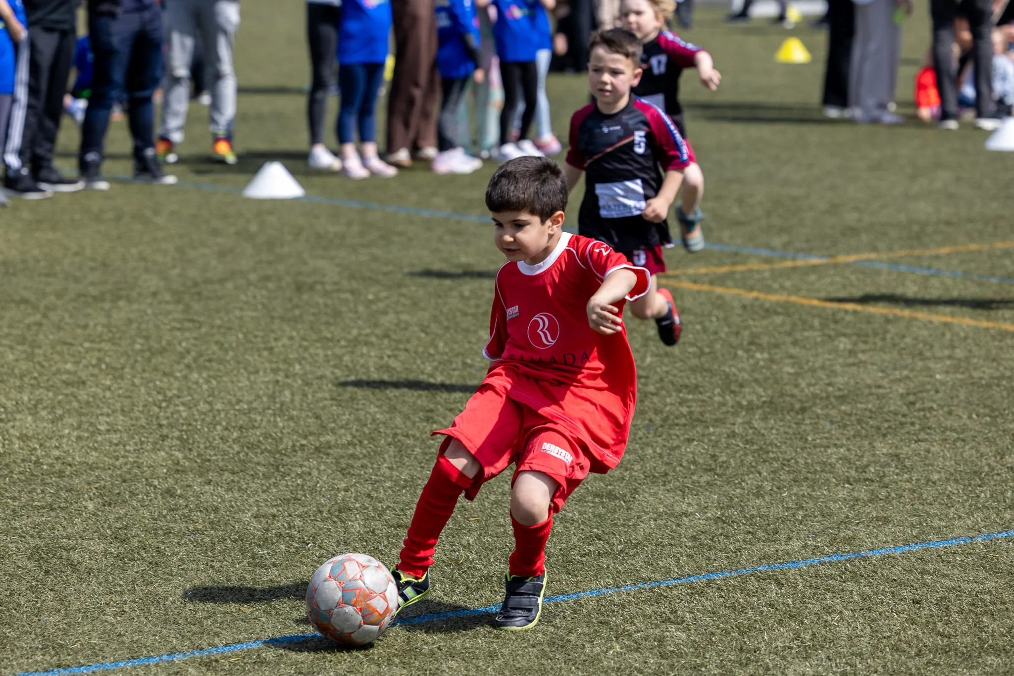 Ein Junge in rotem Fußballtrikot dribbelt den Ball auf einem grünen Rasenfeld, während andere Kinder im Hintergrund zusehen.