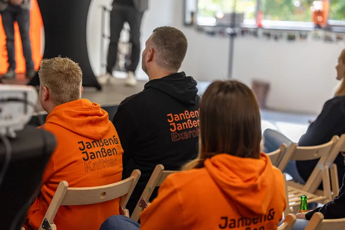 Rückansicht von Personen in orangefarbenen und schwarzen Kapuzenpullovern mit dem Aufdruck „Janßen&Janßen“.