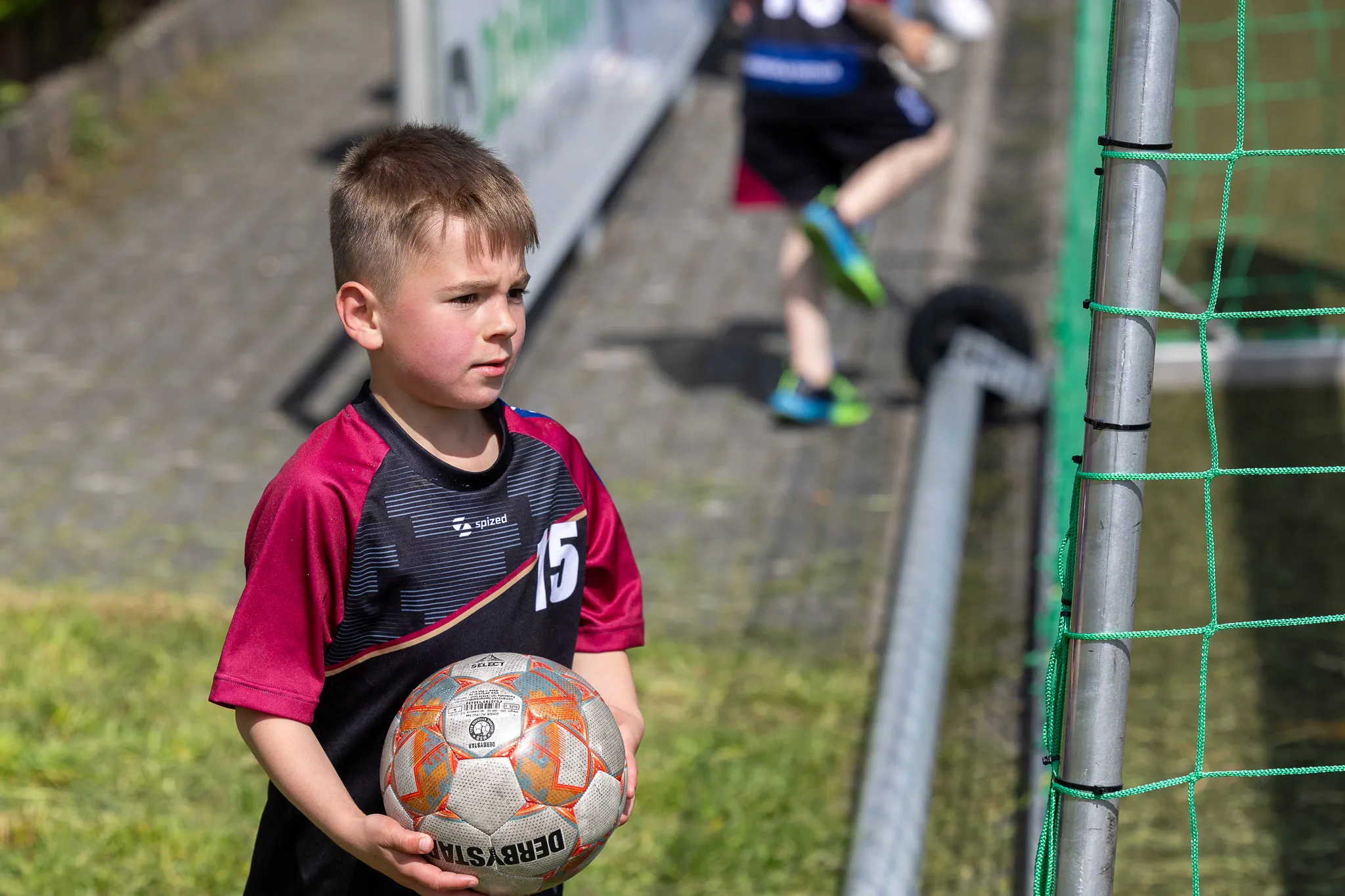 Ein Junge hält einen Fußball vor sich, während im Hintergrund andere Kinder Fußball spielen. Das Turnier findet am Kreisch statt.