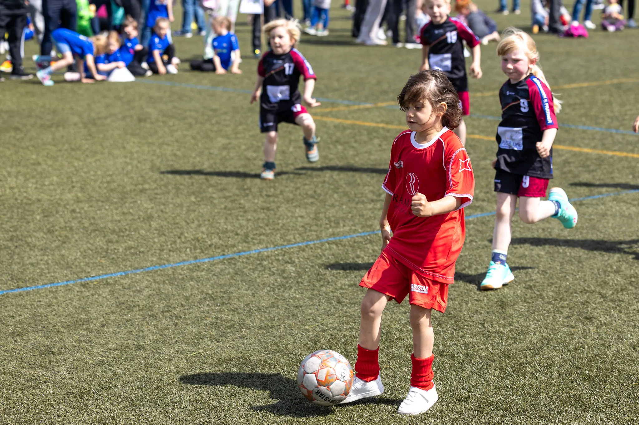 Ein junges Mädchen in rotem Fußballtrikot dribbelt den Ball auf einem Kunstrasenplatz, während andere Kinder im Hintergrund spielen. Das Bild zeigt eine Szene vom Kita-Fußballturnier am Kreisch.