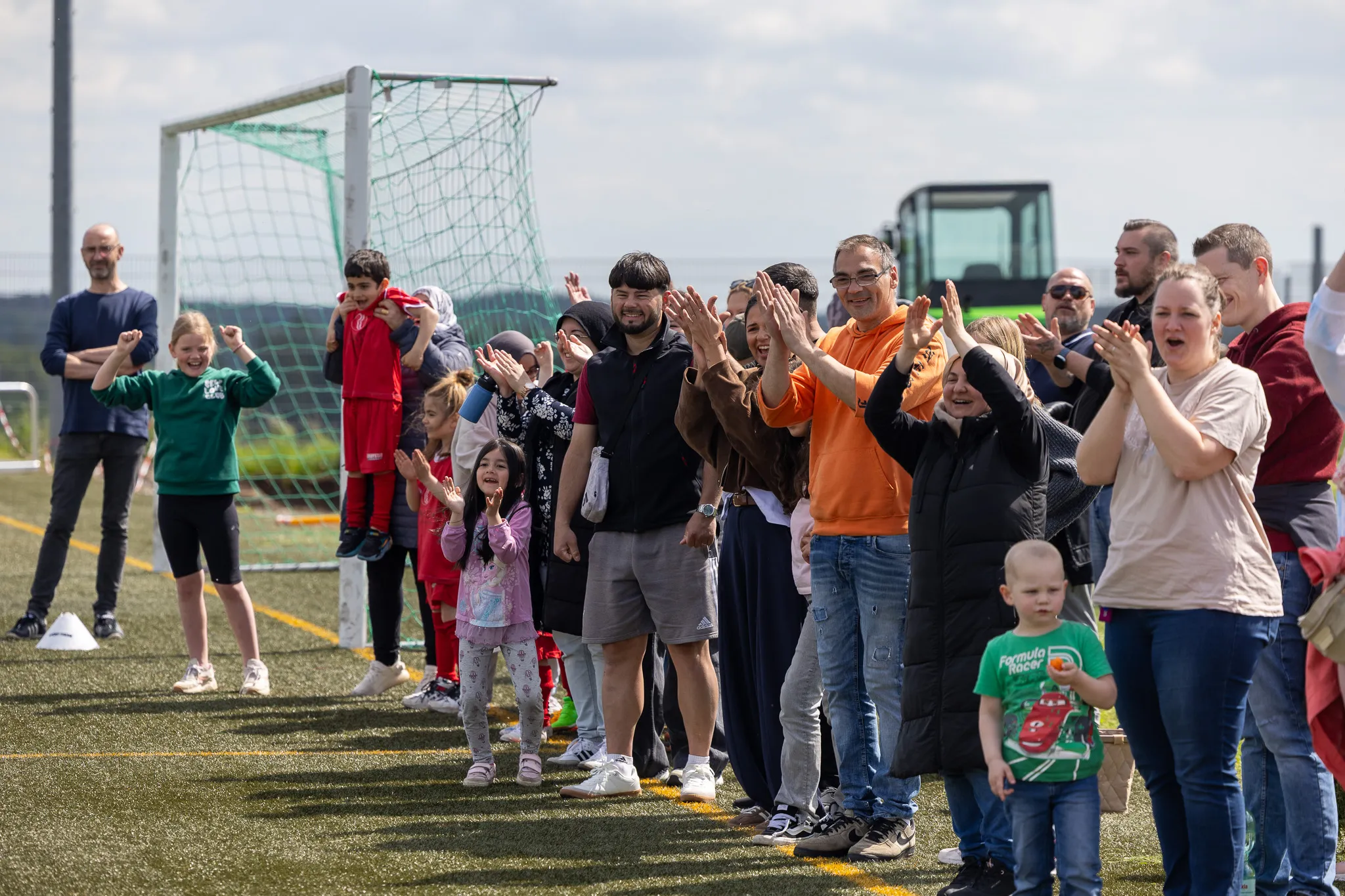 Zuschauer, darunter viele Kinder, klatschen und jubeln an einem Fußballfeld. Im Hintergrund ein Fußballtor und ein Traktor.