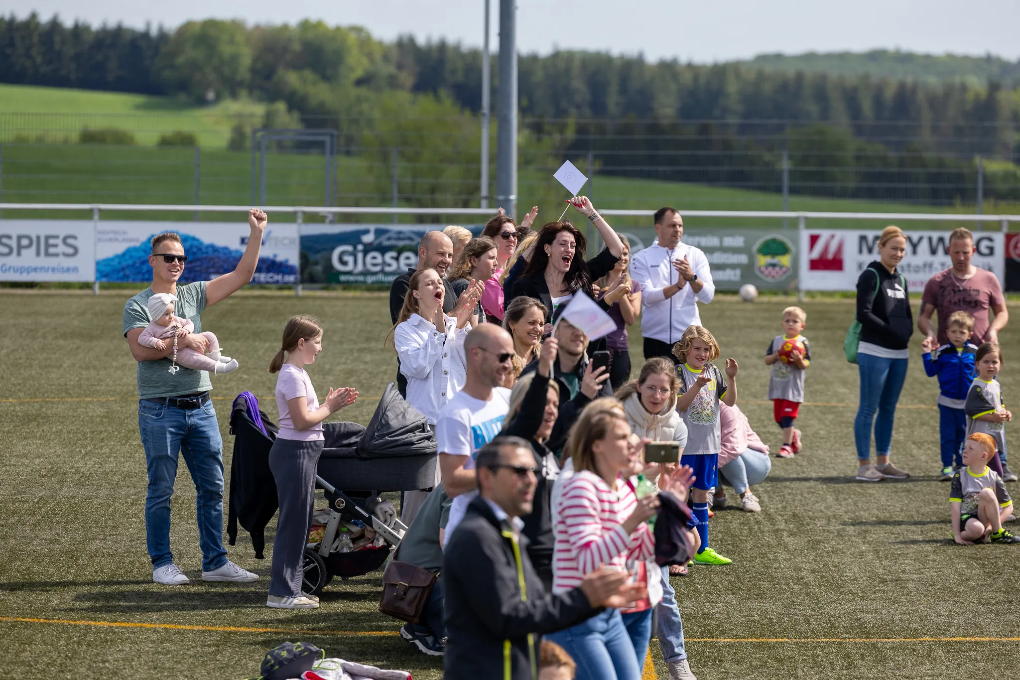 Zuschauer, darunter viele Eltern mit Kindern, jubeln und klatschen bei einem Fußballturnier am Kreisch. Im Hintergrund eine grüne Landschaft und Werbebanner.
