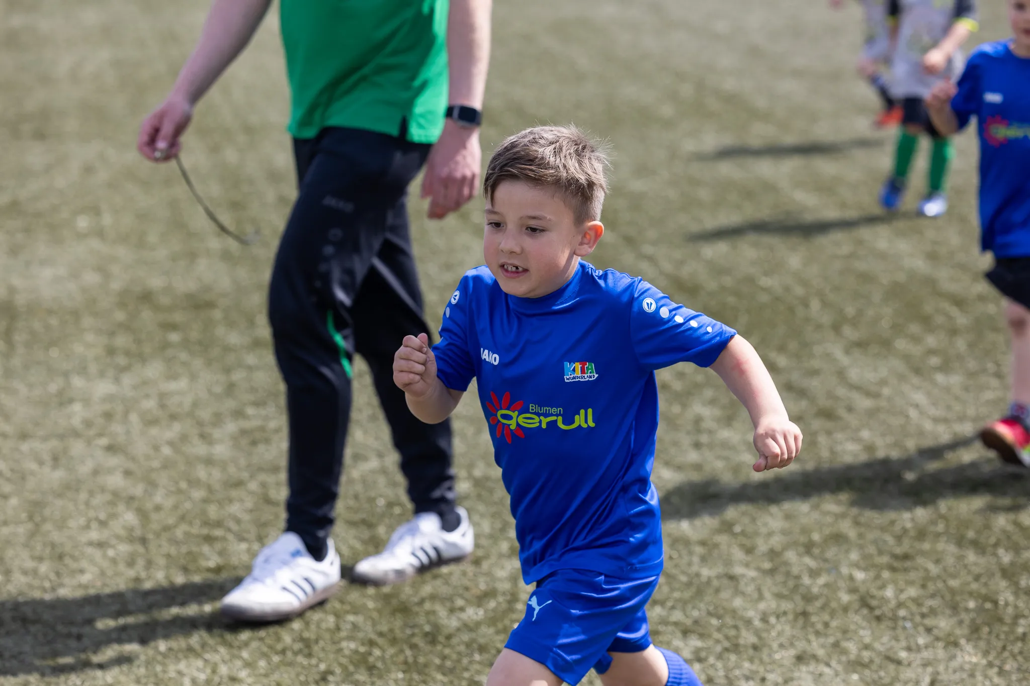 Ein junger Fußballspieler in blauem Trikot rennt auf dem Spielfeld, während im Hintergrund weitere Kinder und Betreuer zu sehen sind.