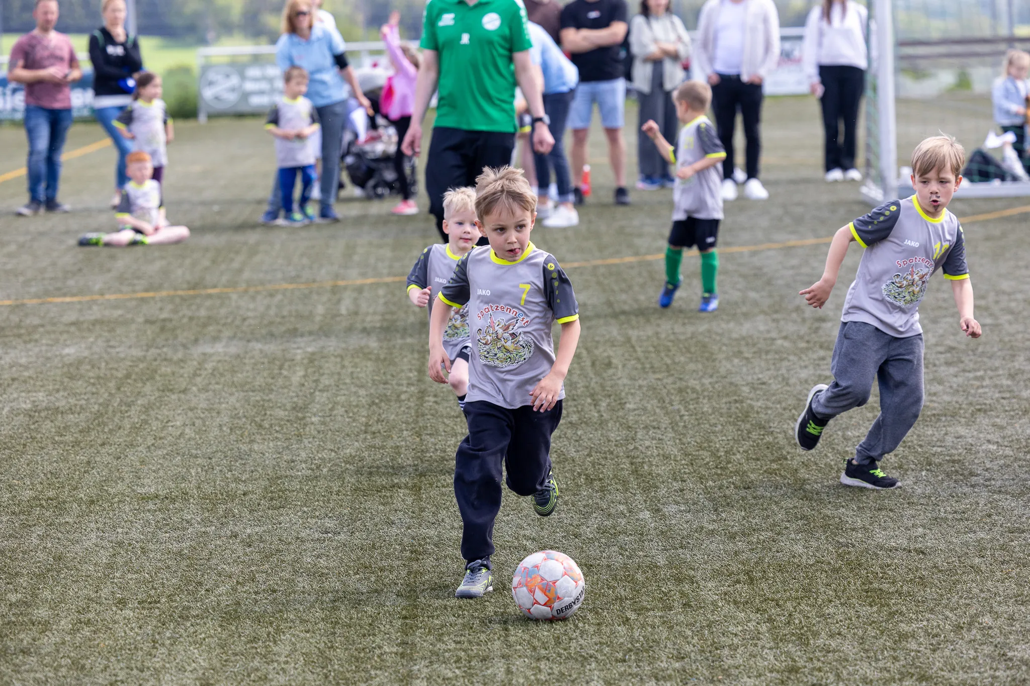 Zwei kleine Fußballspieler in Trikots rennen auf einen Ball zu, während im Hintergrund Zuschauer stehen.