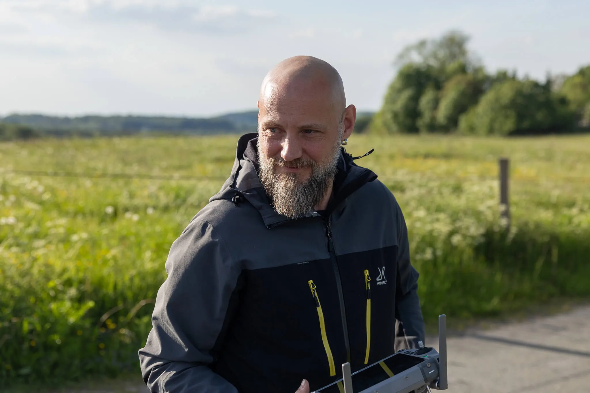 Ein Mann mit Glatze und Bart lächelt in die Kamera, während er eine Drohne hält. Im Hintergrund eine ländliche Landschaft mit Feldern und Bäumen.