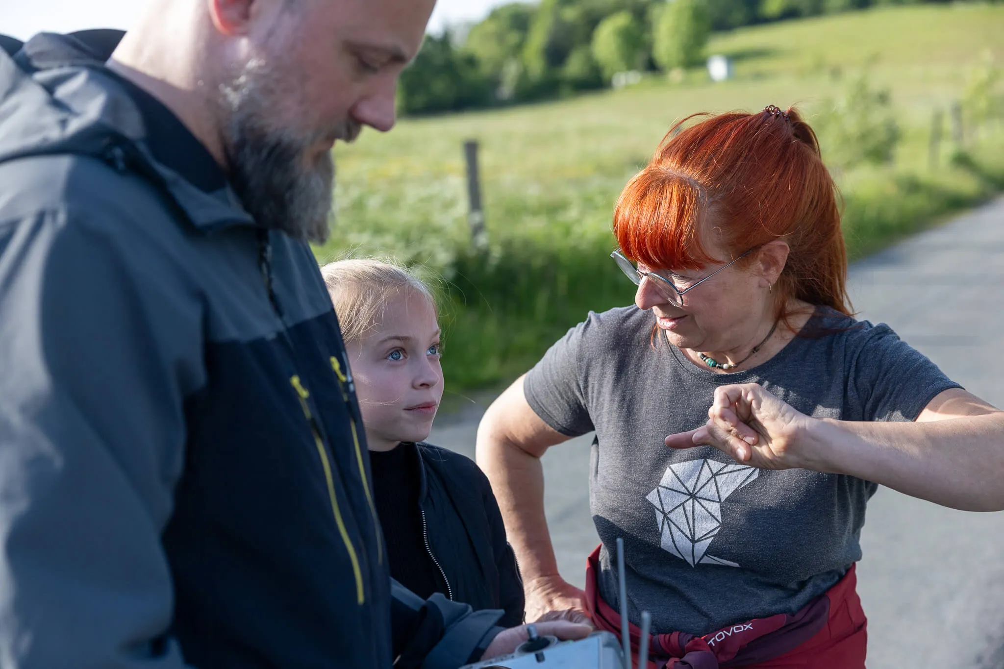 Eine Frau mit roten Haaren erklärt einem Mann und einem Kind etwas, während sie auf eine Fernbedienung zeigt. Im Hintergrund eine grüne Landschaft.