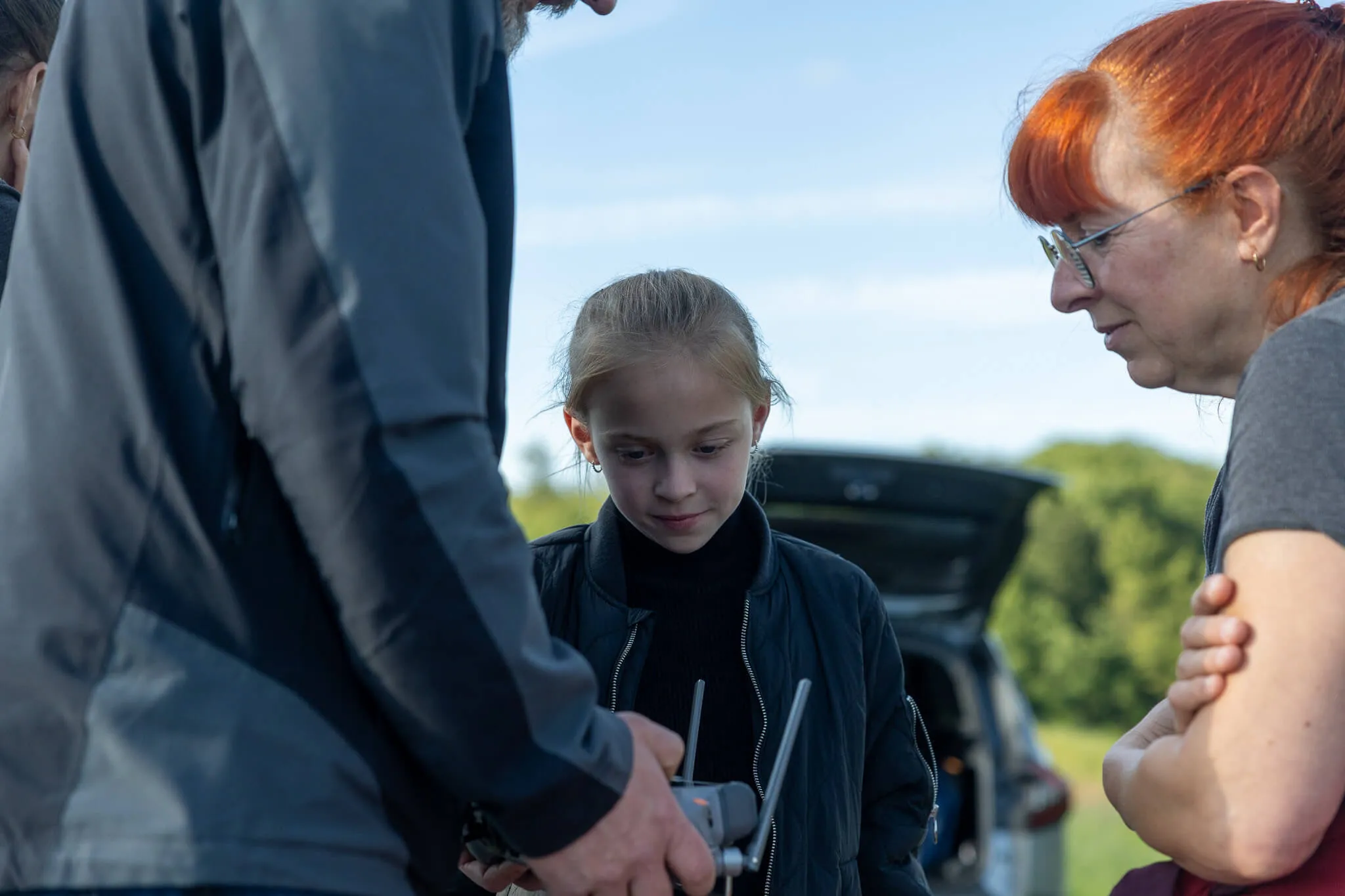 Ein Mädchen blickt auf ein ferngesteuertes Gerät, das ein Mann hält, während eine Frau im Hintergrund beobachtet. Im Hintergrund ist ein geöffneter Kofferraum eines Autos zu sehen.