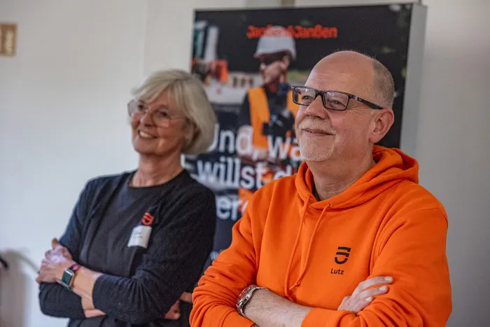 Ein lächelnder Mann in einem orangefarbenen Kapuzenpullover und eine Frau mit grauen Haaren stehen vor einem Poster der Firma Janßen & Janßen.