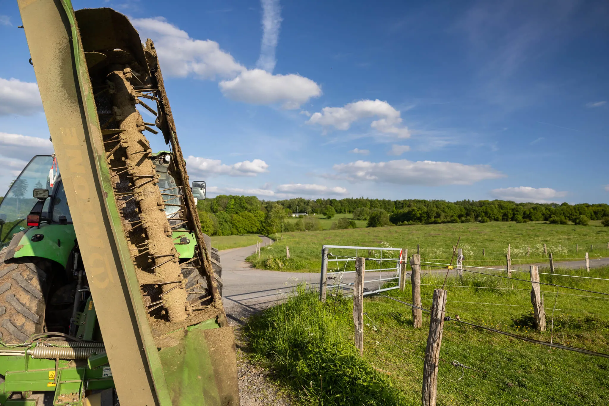 Ein Traktor mit einem angebauten Gerät zum Aufnehmen von Gras oder Heu steht auf einem Feldweg. Im Hintergrund erstreckt sich eine grüne Landschaft mit Bäumen und einem blauen Himmel.
