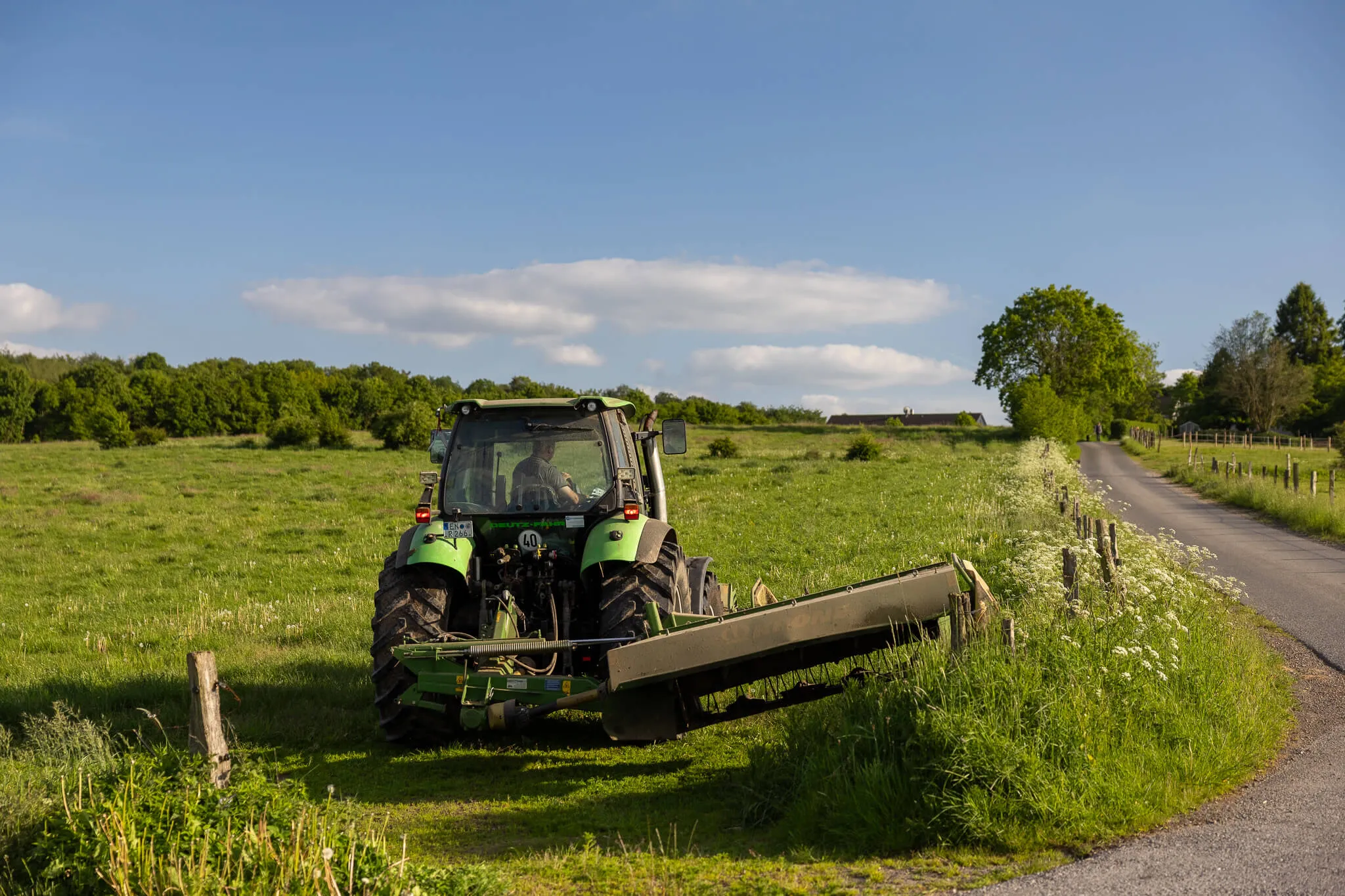 Grüner Traktor mit Mähwerk fährt an einem Feldweg entlang, während ein Mann im Fahrersitz sitzt. Im Hintergrund eine ländliche Landschaft unter blauem Himmel.