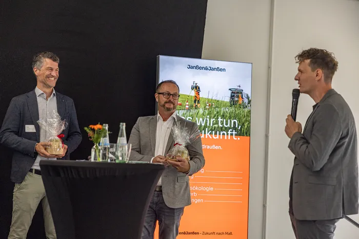 Zwei Männer halten Geschenke vor einem Janßen&Janßen-Banner, während ein dritter Mann mit einem Mikrofon zu ihnen spricht.