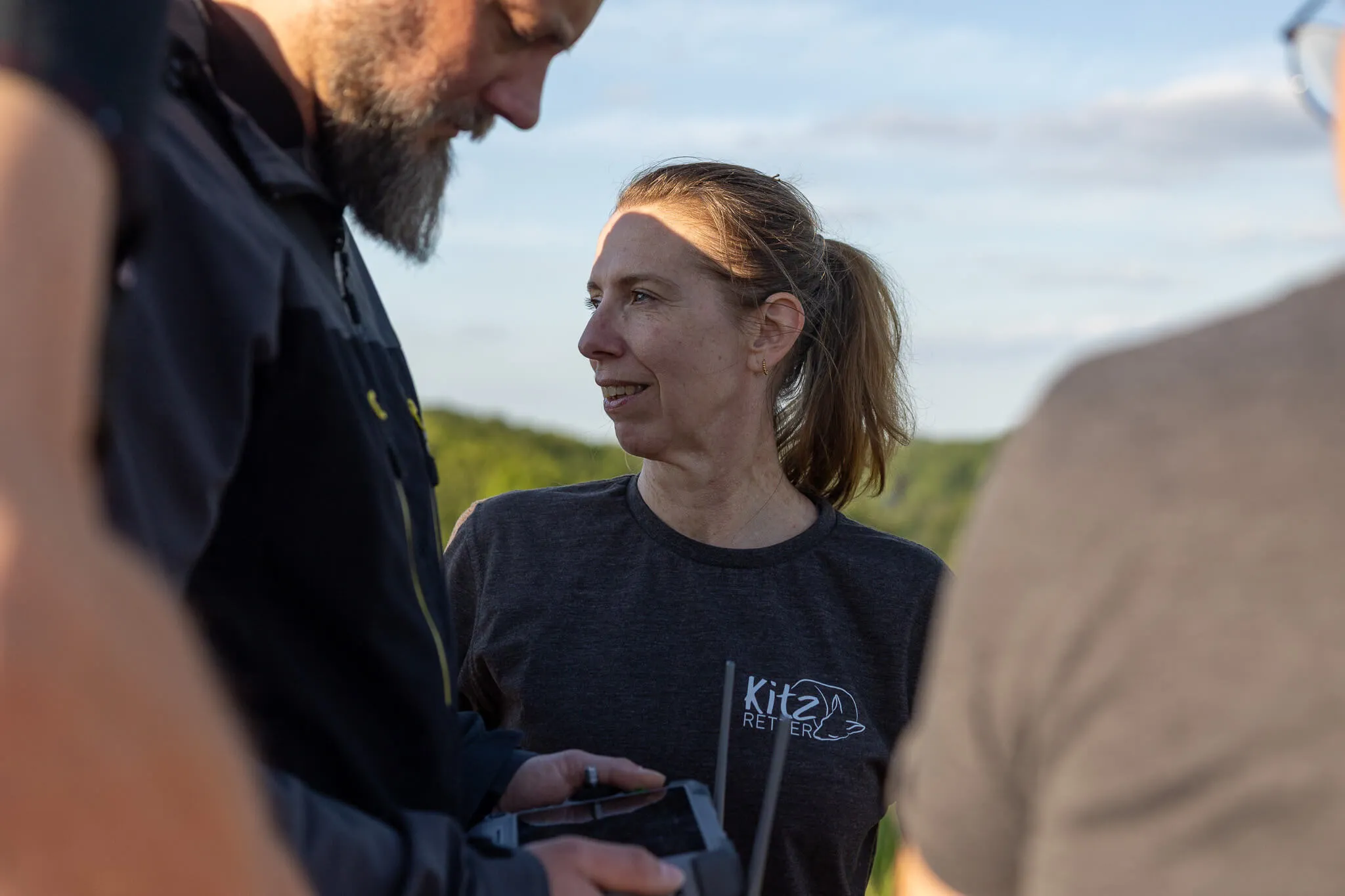Eine Frau mit hochgestecktem Haar lächelt, während sie mit zwei Männern im Freien spricht. Sie trägt ein T-Shirt mit dem Aufdruck „Kitz RETT ER“.
