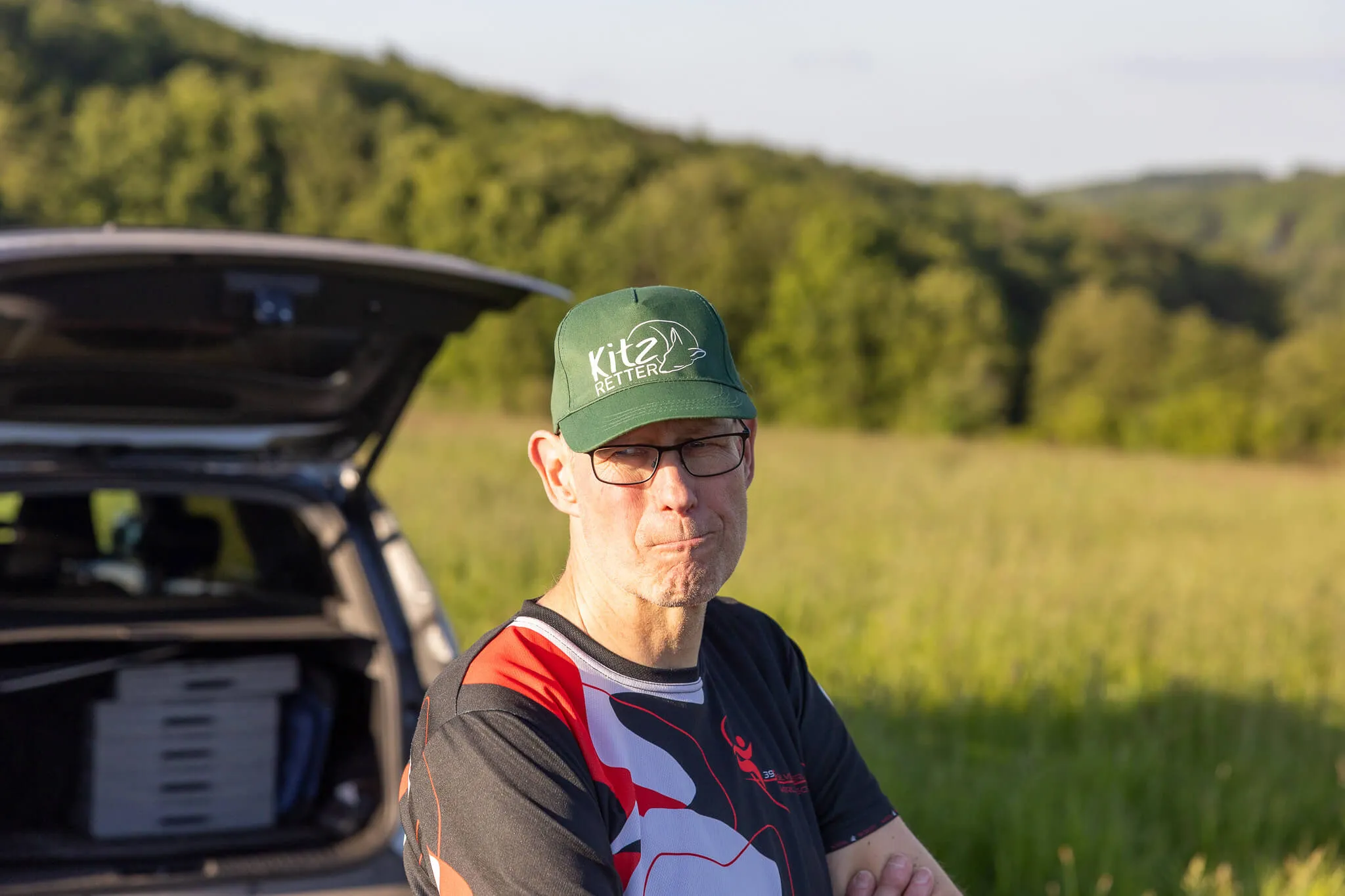Ein Mann mit Brille und Kappe steht vor einem geöffneten Kofferraum eines Autos auf einer Wiese. Im Hintergrund sind Bäume zu sehen.