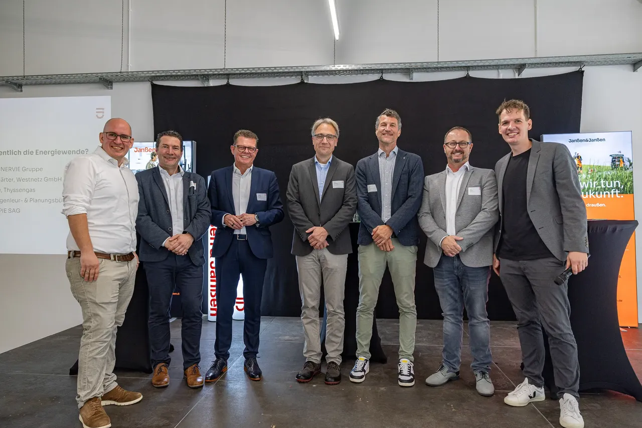 Sieben Männer posieren für ein Gruppenfoto vor einem schwarzen Hintergrund bei der Jubiläumsfeier von Janßen & Janßen.