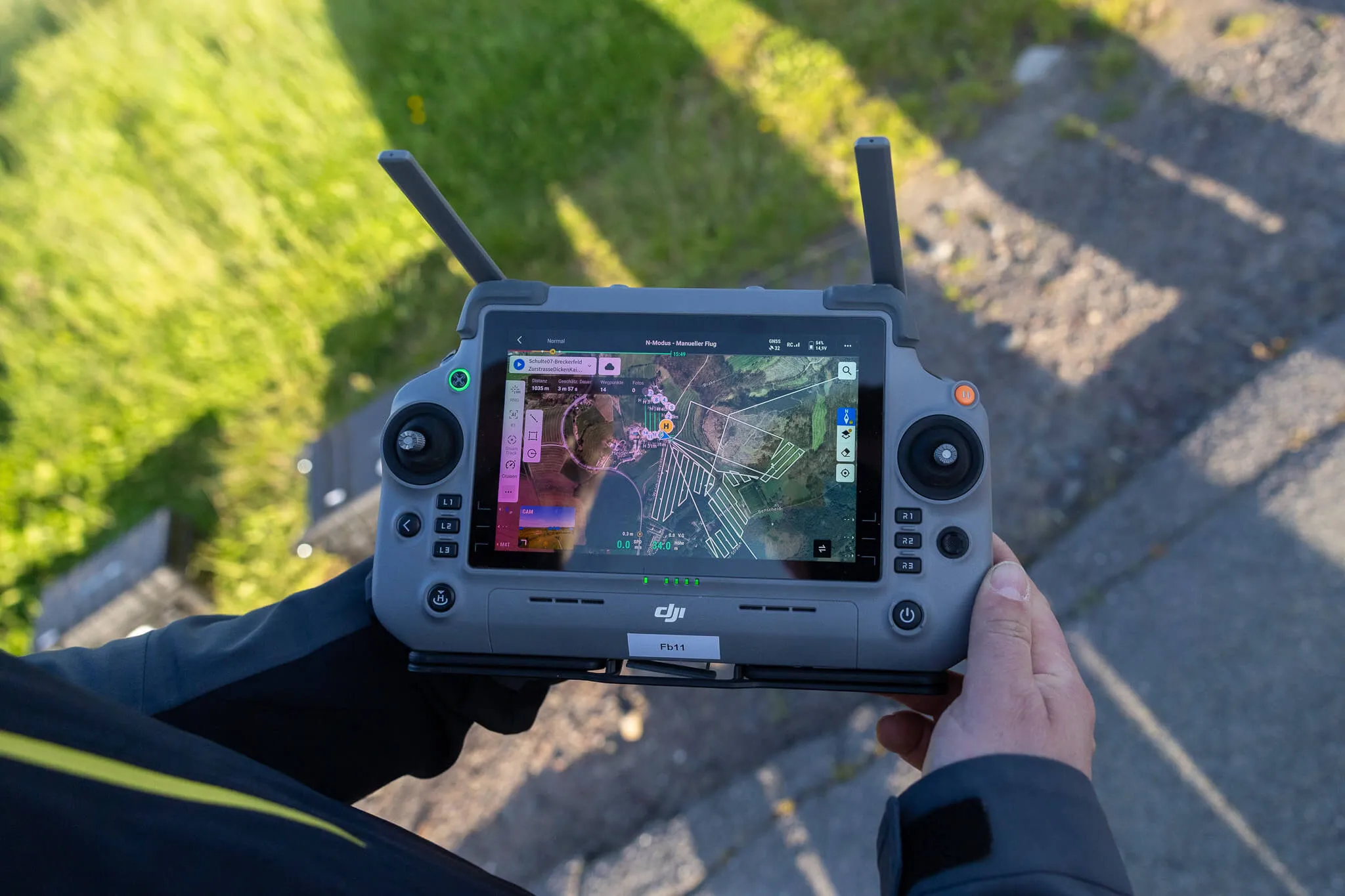 Hände halten einen DJI-Fernbedienung mit einem hellen Bildschirm, der eine Karte und Flugdaten anzeigt. Im Hintergrund ist grünes Gras und eine Steinmauer zu sehen.