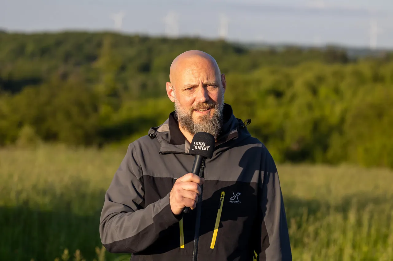 Mann mit Bart hält Mikrofon im Feld, im Hintergrund Bäume und Windräder. Er trägt eine Jacke mit dem Aufdruck „Lokal Direkt“.