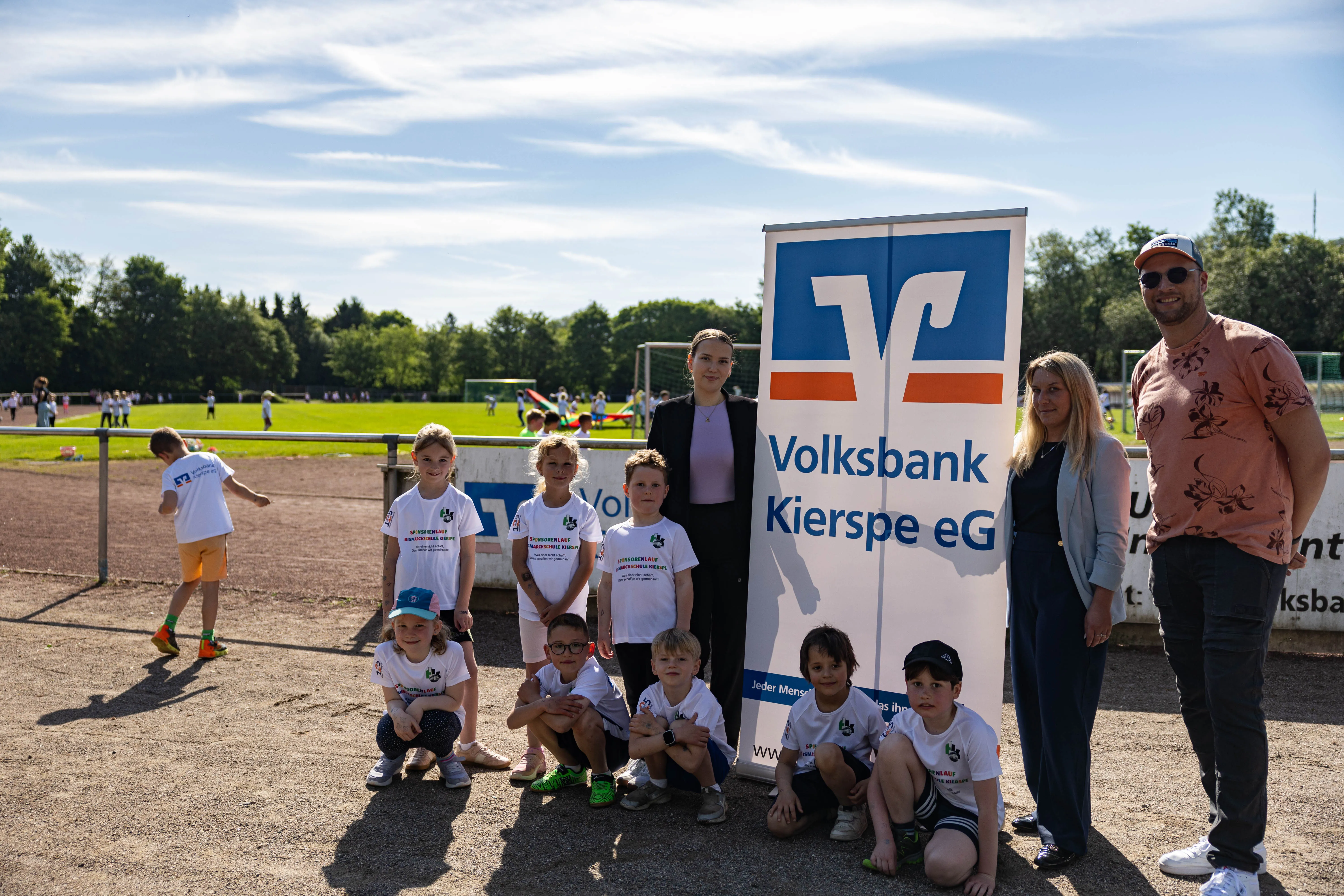 Gruppe von Kindern in Sportkleidung und zwei Erwachsene vor einem Werbebanner der Volksbank Kierspe eG während eines Spendenlaufs.