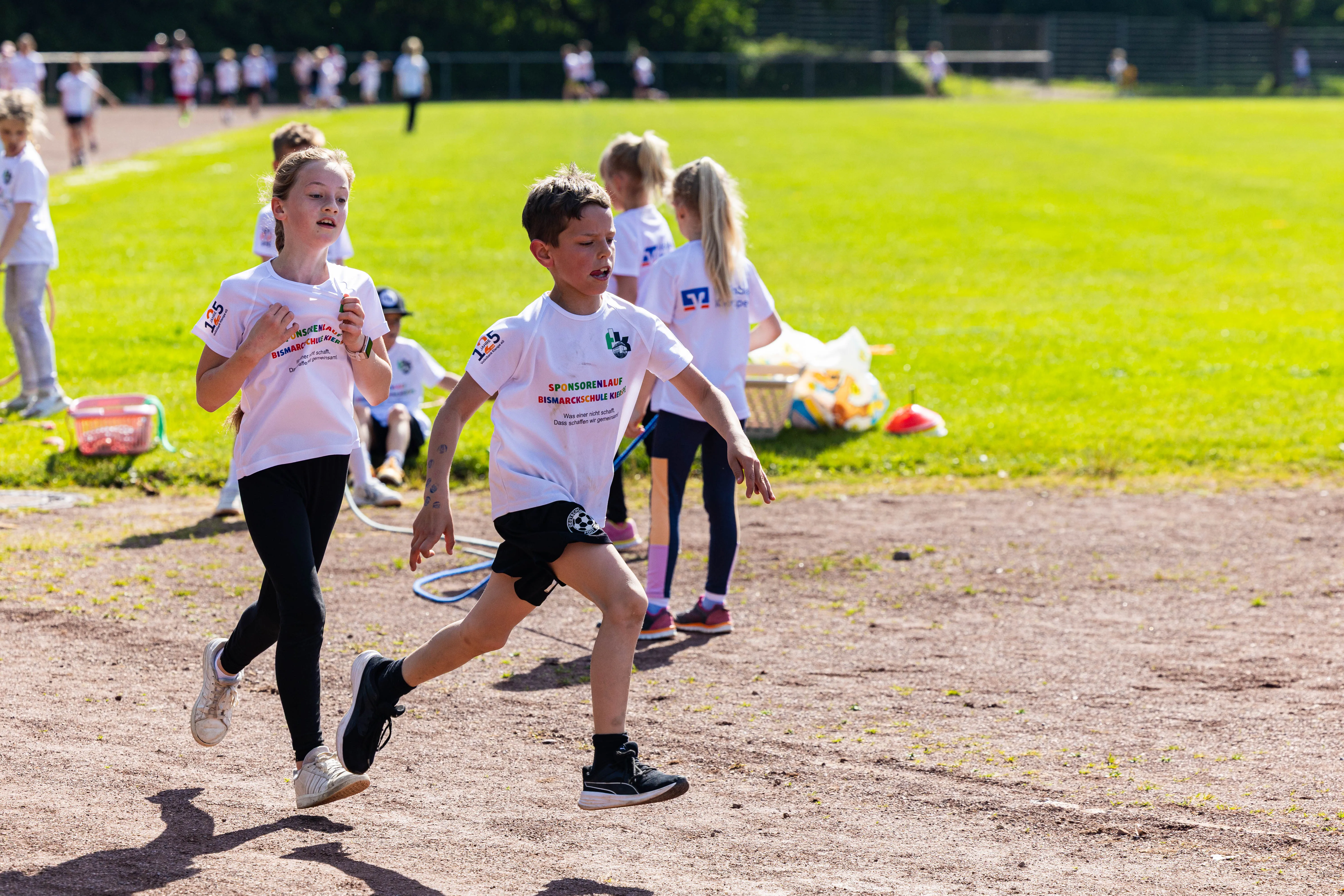 Zwei Kinder laufen beim Spendenlauf der Bismarckschule Kierspe auf einer Tartanbahn. Im Hintergrund weitere Kinder und ein grüner Rasen.