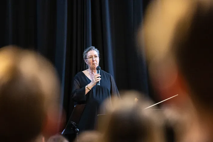 Eine Frau mit grauen Haaren und Brille spricht in ein Mikrofon auf einer Bühne. Im Hintergrund sind verschwommene Köpfe von Zuhörern und ein schwarzer Vorhang zu sehen.