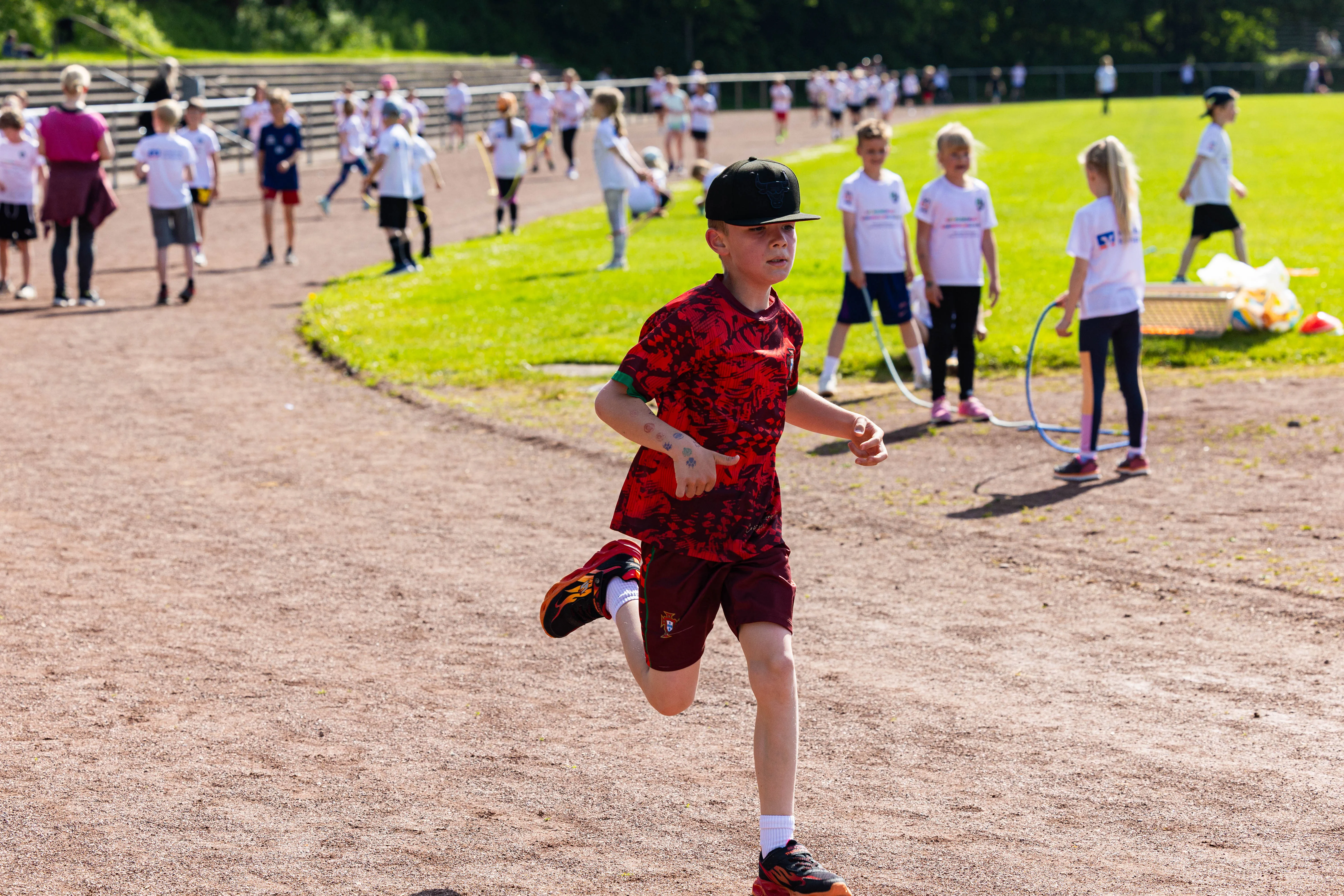 Ein Junge mit Baseballkappe rennt auf einer roten Tartanbahn, im Hintergrund weitere Kinder und Zuschauerränge.