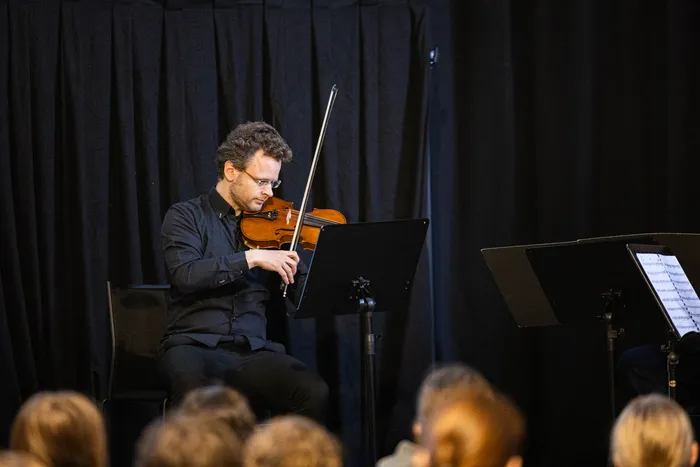 Ein Mann spielt Violine vor Notenständern und einem Publikum. Er sitzt auf einem Stuhl und trägt ein schwarzes Hemd. Im Hintergrund sind dunkle Vorhänge zu sehen.