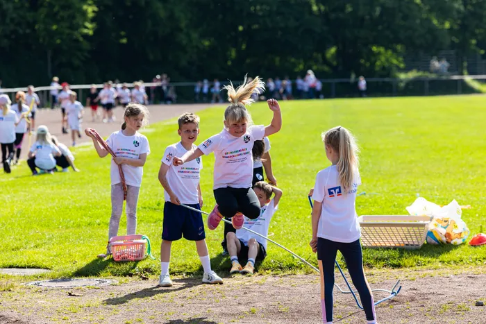 Schülerinnen und Schüler der Bismarckschule Kierspe laufen bei einem Spendenlauf auf einer grünen Wiese, einige mit roten und blauen Körben. Ein Mädchen springt über ein Hindernis.