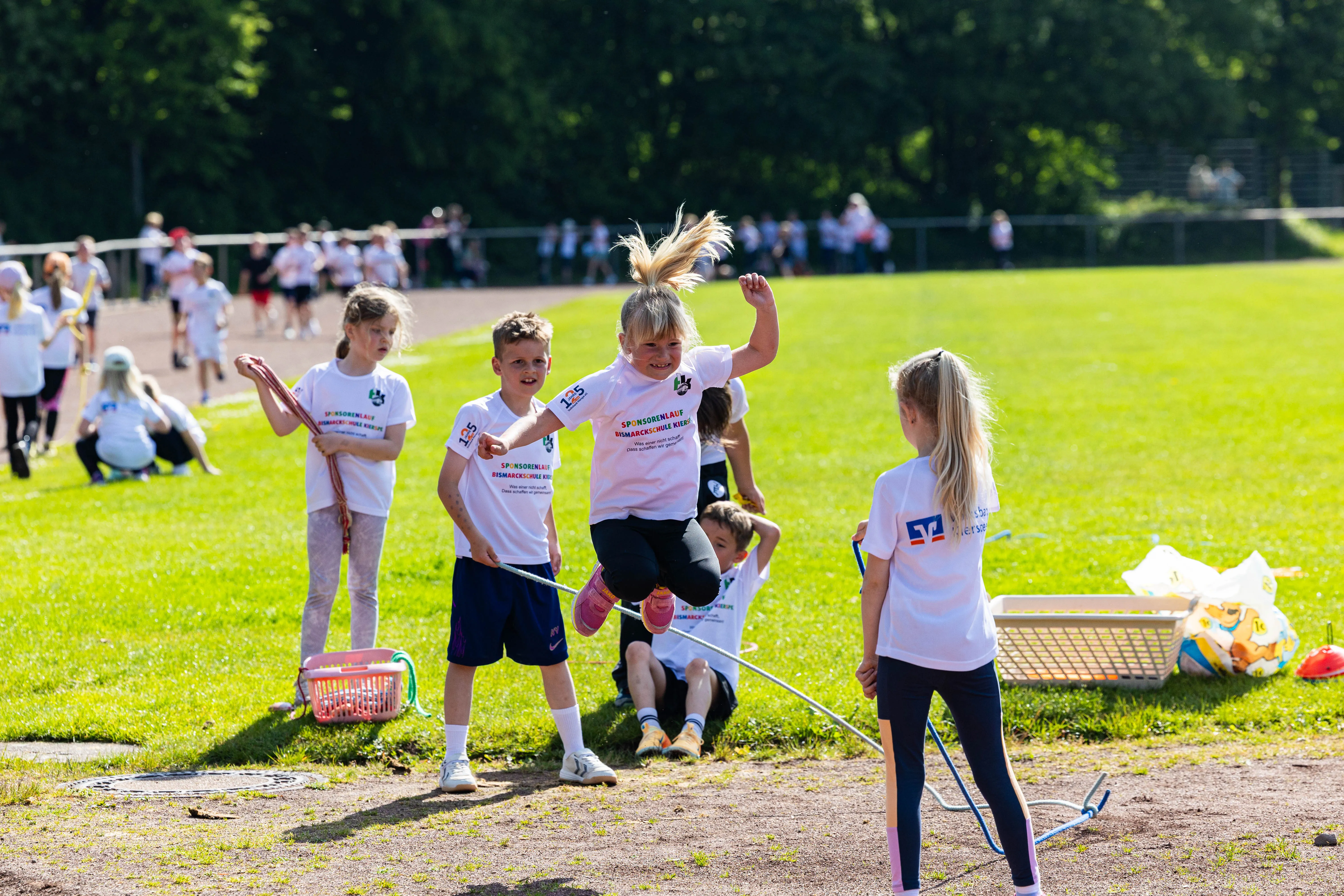 Schülerinnen und Schüler der Bismarckschule Kierspe laufen bei einem Spendenlauf auf einer grünen Wiese, einige mit roten und blauen Körben. Ein Mädchen springt über ein Hindernis.