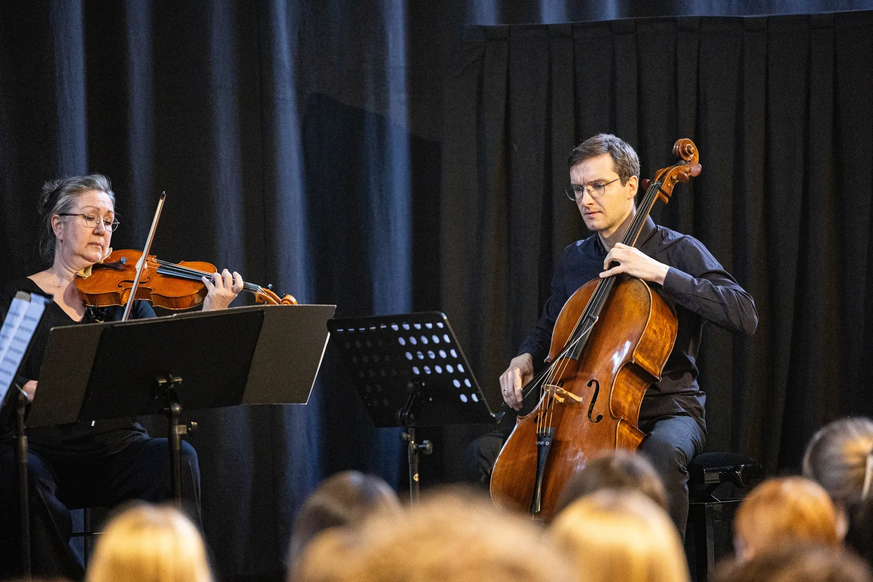 Ein Geiger und ein Cellist spielen während eines Konzerts vor einem dunklen Hintergrund. Beide Musiker sitzen auf Stühlen und konzentrieren sich auf ihre Notenblätter. Im Hintergrund sind Köpfe von Zuschauern zu sehen.