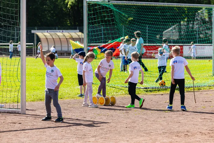 Kindergruppe läuft auf einem Fußballfeld, einige halten gelbe Spendenbehälter. Im Hintergrund ein Fußballtor und ein Vereinsgebäude.