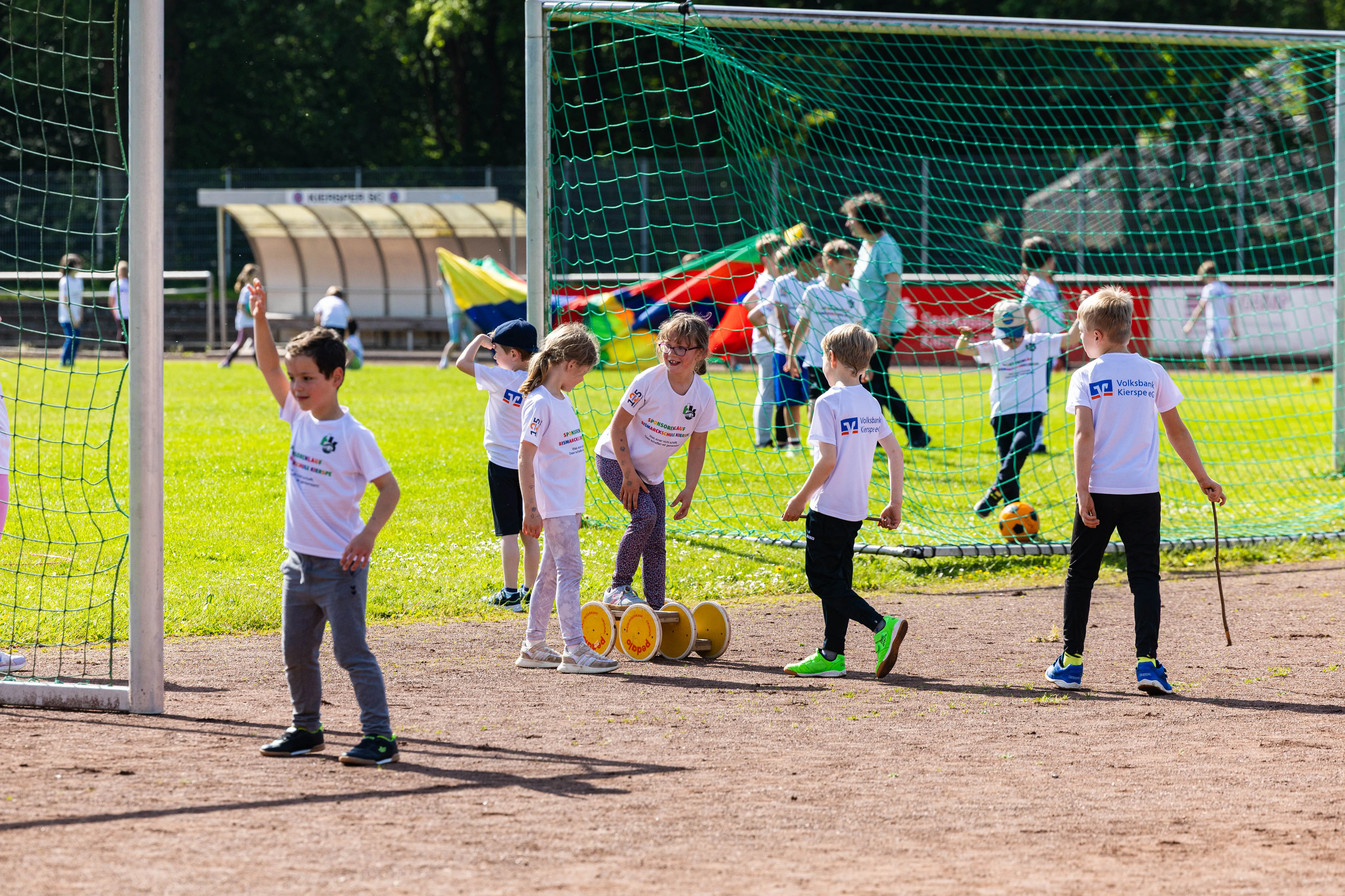 Kindergruppe läuft auf einem Fußballfeld, einige halten gelbe Spendenbehälter. Im Hintergrund ein Fußballtor und ein Vereinsgebäude.