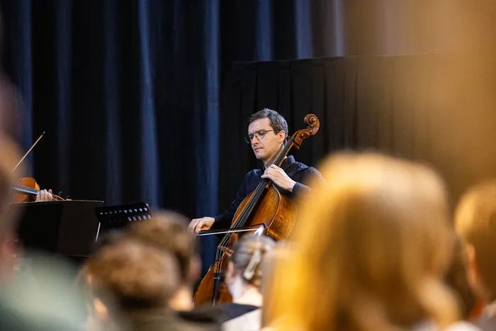 Ein Cellist spielt während eines Konzerts im Kulturort 8Giebel. Im Hintergrund sind weitere Musiker und das Publikum zu erkennen.