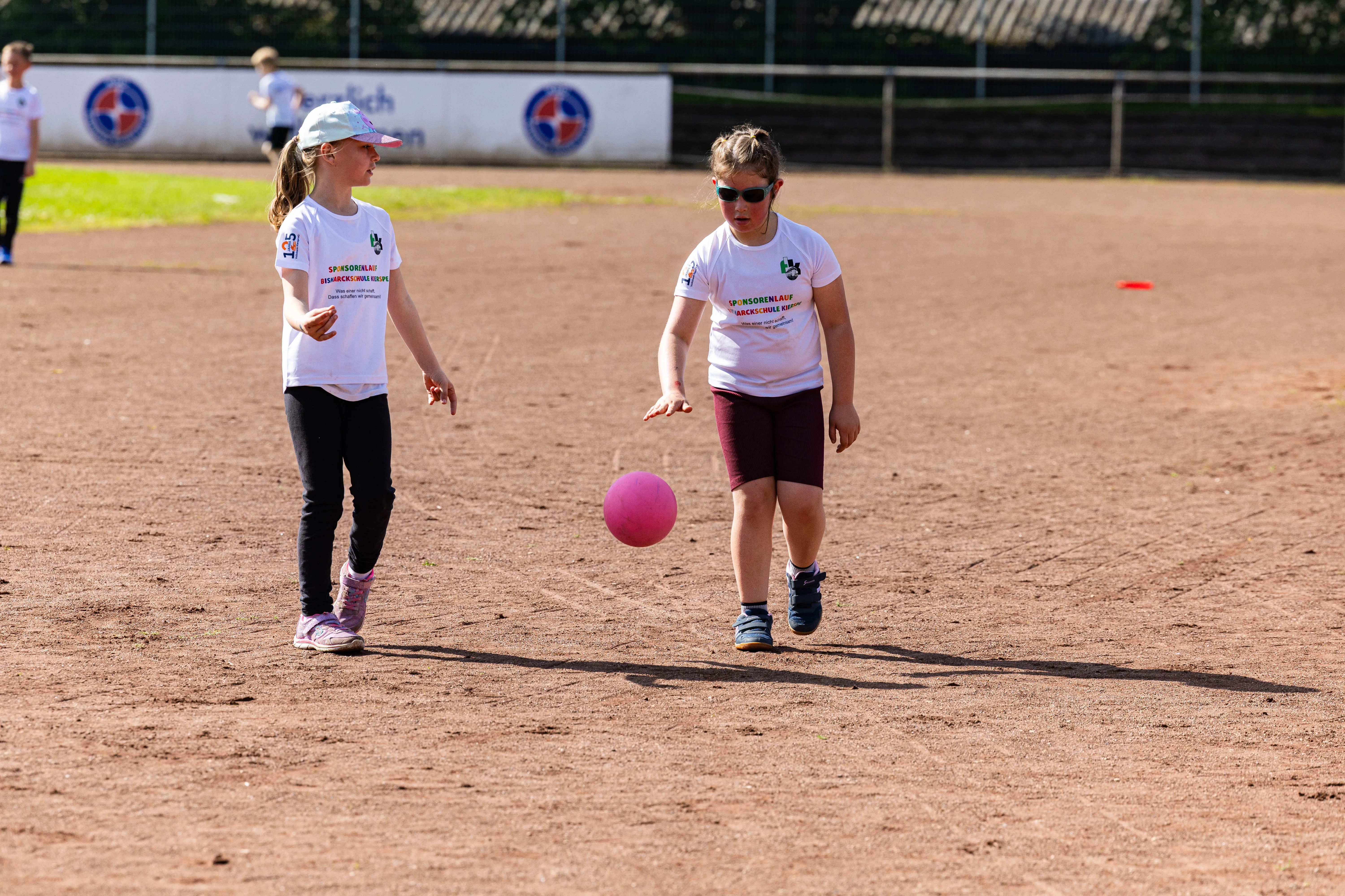 Zwei Schülerinnen mit T-Shirts und Sonnenbrillen laufen auf einer roten Tartanbahn, eine hält einen rosa Ball.